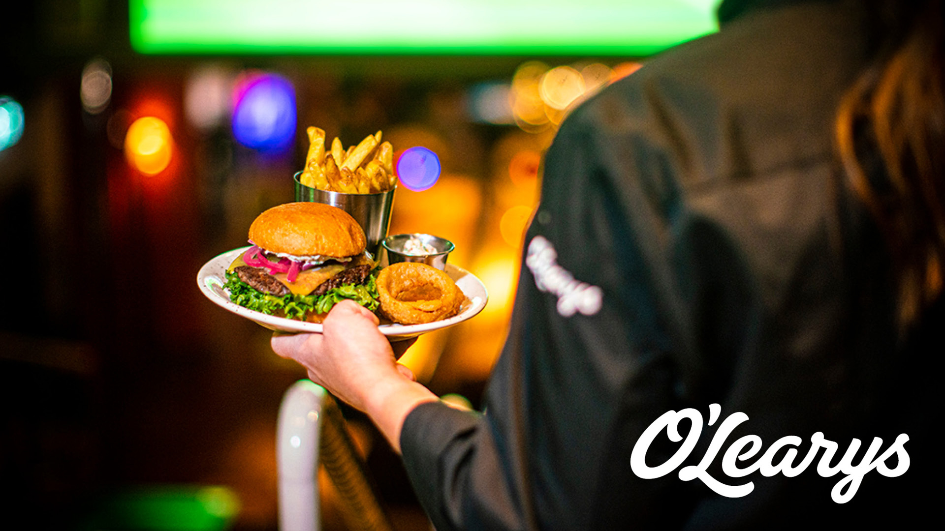 Servitör på O'Learys med en tallrik innehållande hamburgare, pommes frites och lökringar, mot en livlig och färgglad bakgrund