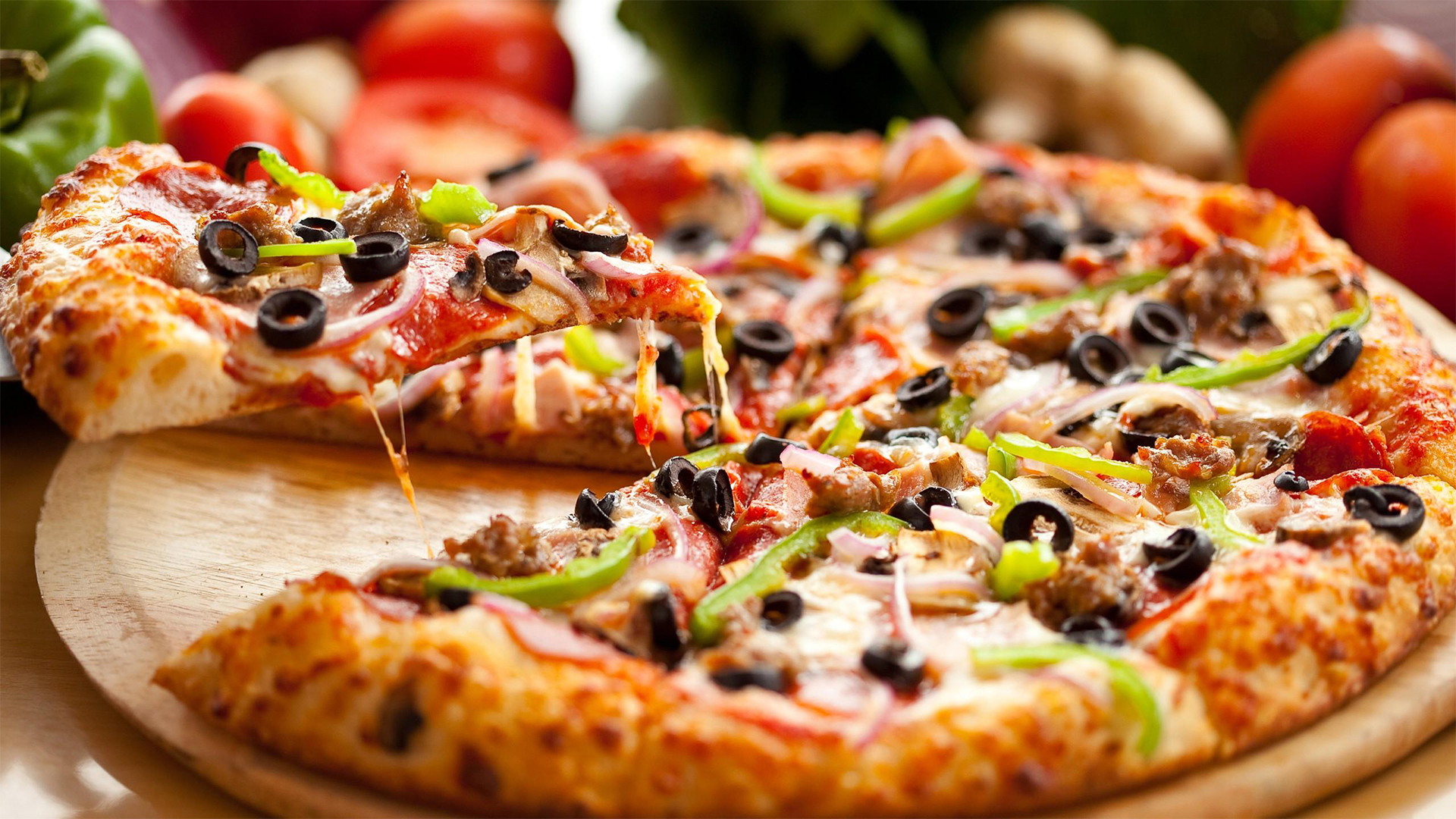 Pizza med ost, skinka, svarta oliver, grön paprika och lök på en bakpanna, omgiven av färska ingredienser