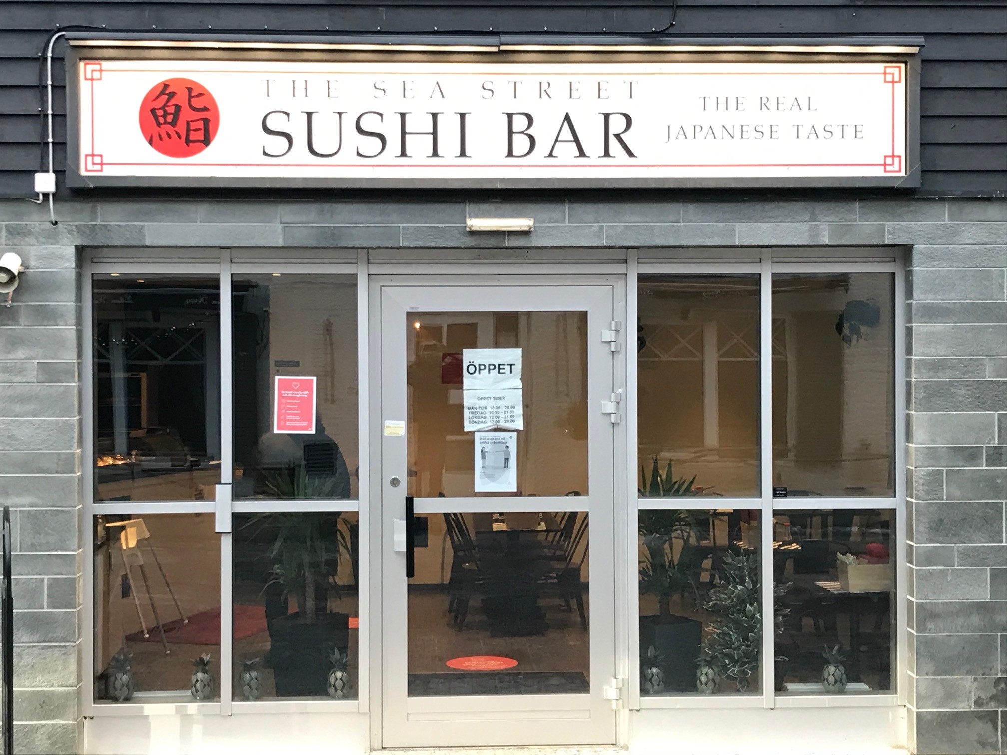 Entré till The Sea Street Sushi Bar med skylt och öppettid på dörren, glasfasad och inredning synlig i bakgrunden