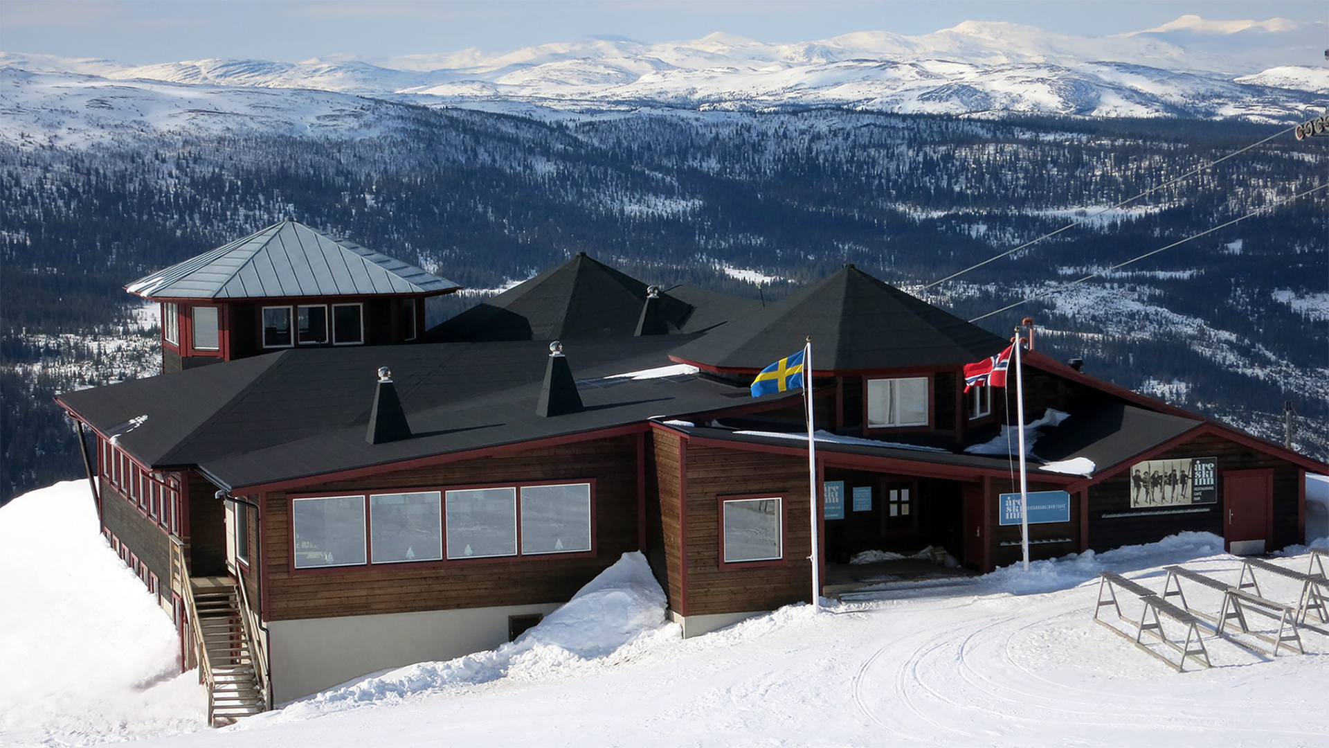 Fjällstuga med svenska och norska flaggor på snötäckt fjäll, omgiven av vinterlandskap