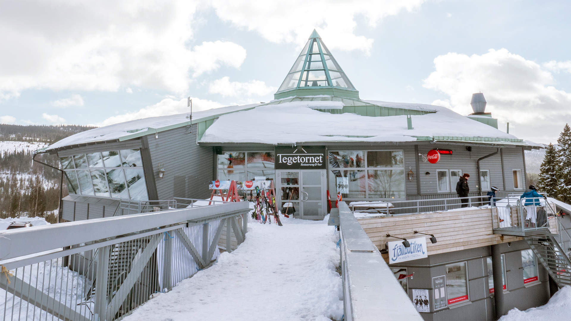 Snötäckt restaurangbyggnad på fjället med skyltar för Toppstugan och Fjällbutiken, en klar himmel i bakgrunden