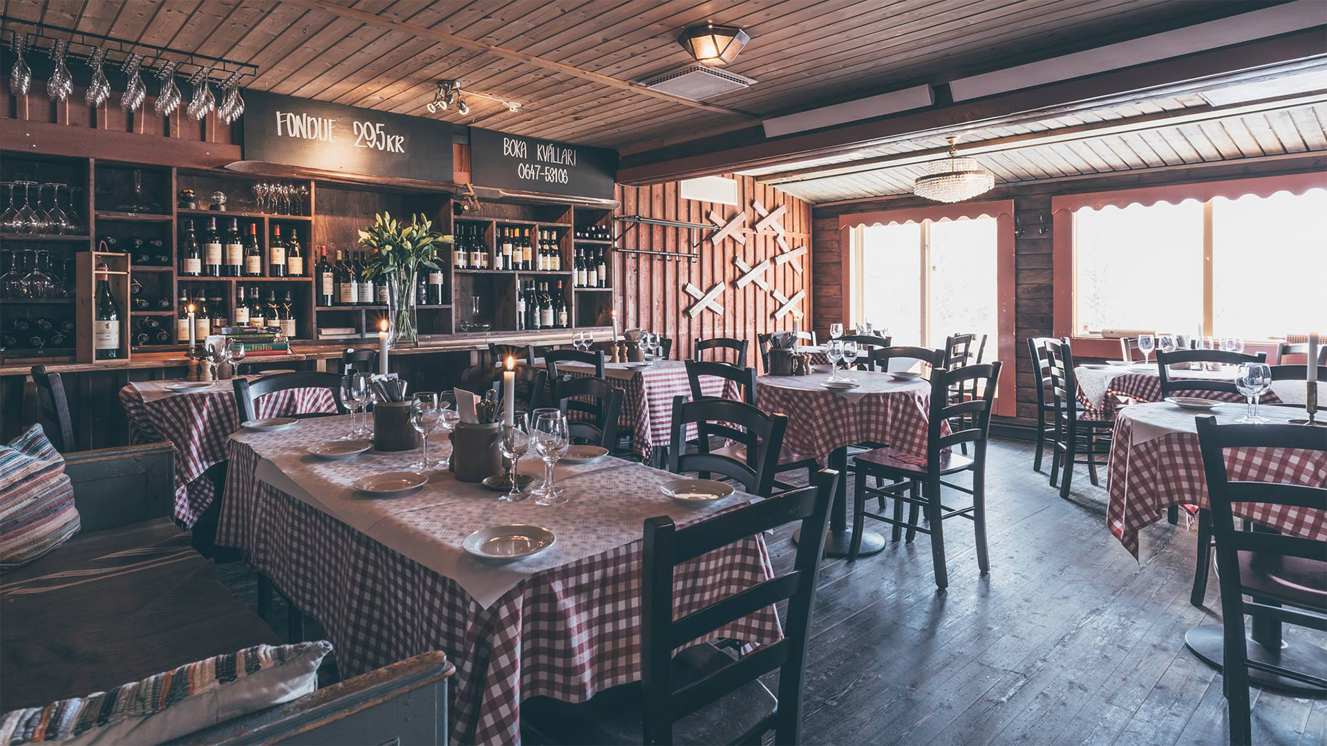Rustik restauranginteriör med röd-vit rutiga dukar, träinredning, vinglas och flaskor. Mysig atmosfär med skyltar som visar fonduepris och bokningsnummer