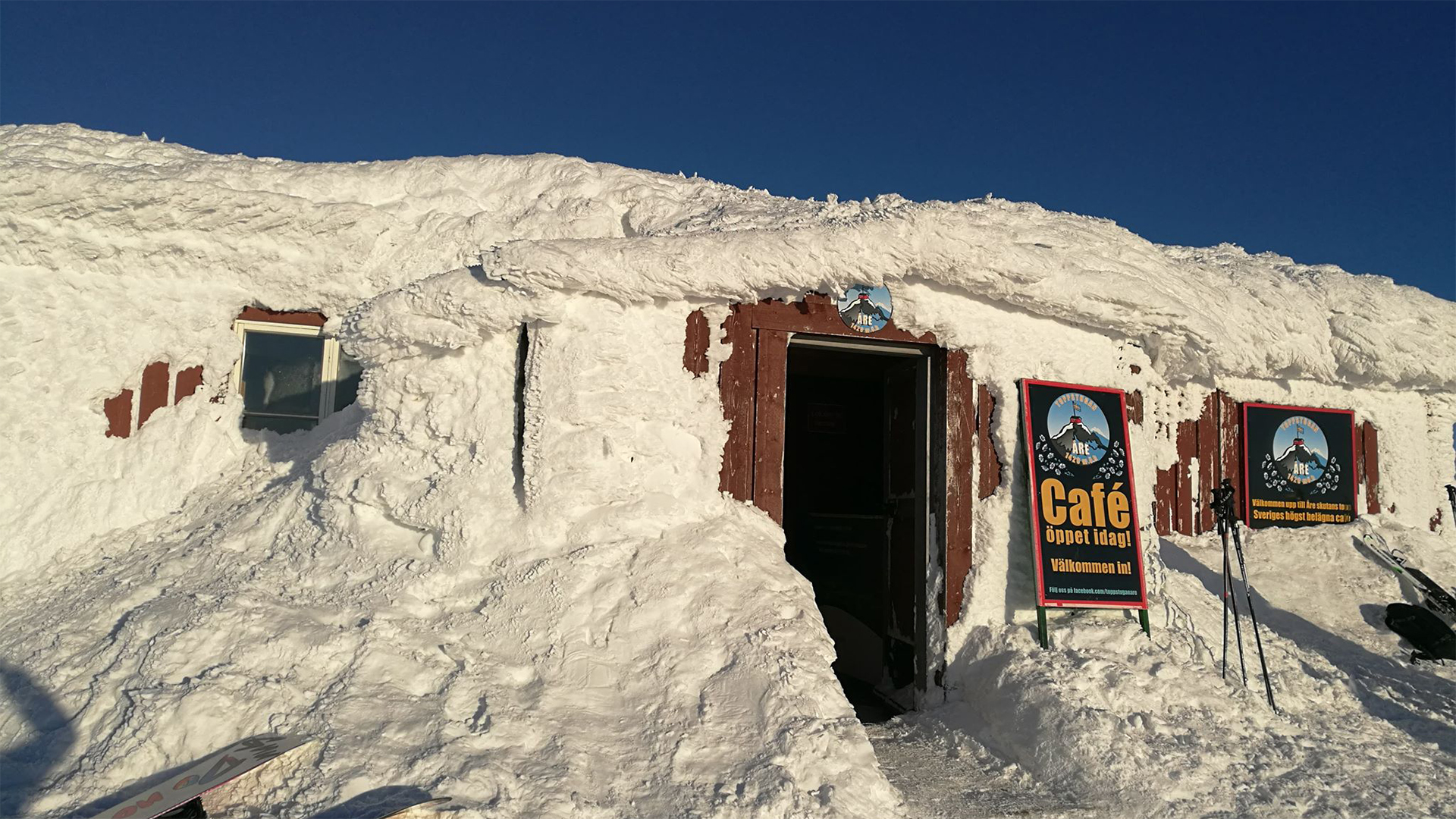 Snötäckt cafébyggnad med skyltar vid ingången, blå himmel i bakgrunden, vinterdag