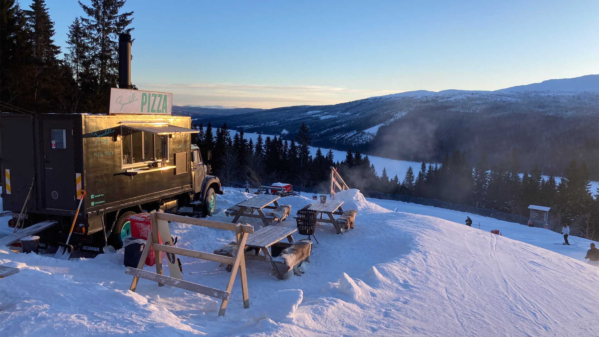 Matvagn som serverar pizza vid en snöig skidbacke i fjällen, med utsikt över vinterlandskap och skog