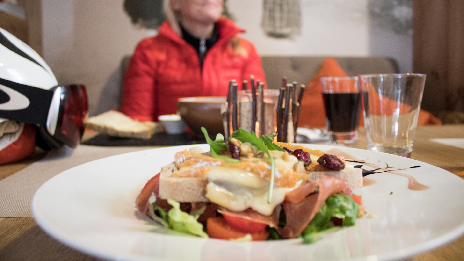 En tallrik med smörgås toppad med ost, skinka och sallad, på ett cafébord. I bakgrunden sitter en person i röd jacka och en cykelhjälm ligger på bordet