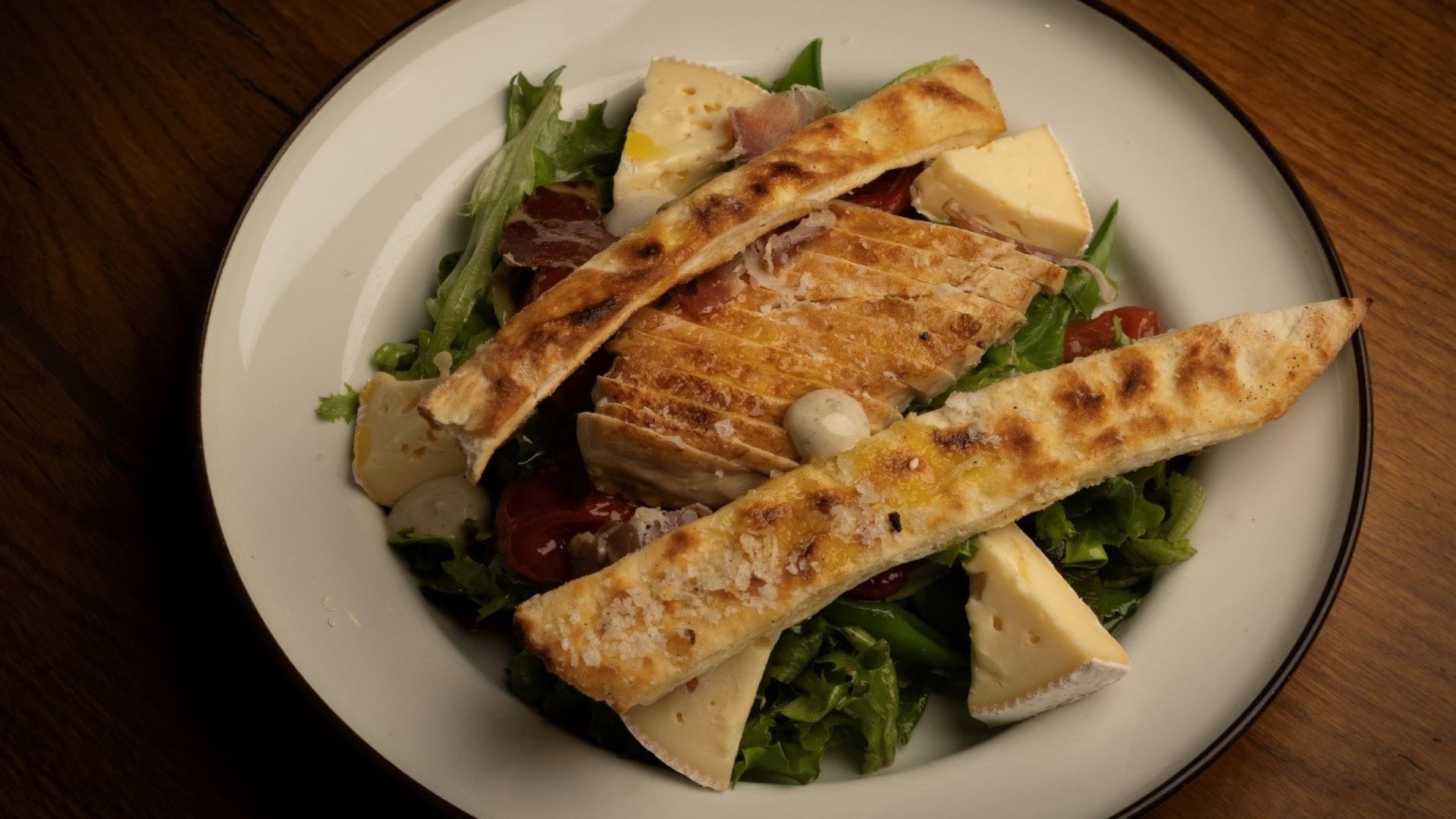 Tallrik med grillad kycklingsallad, brieost, krutonger, parmaskinka och grönsaker på ett träbord