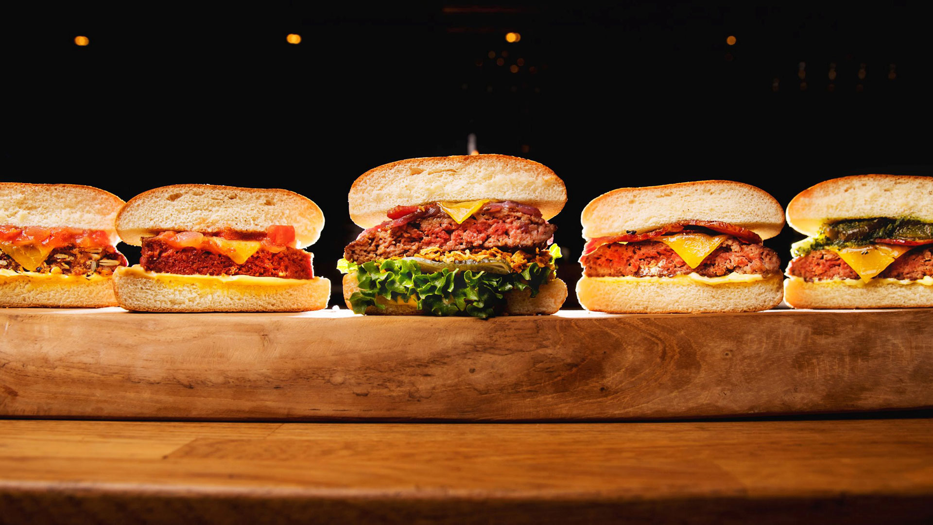 Fem olika hamburgare med varierande fyllningar och pålägg på ett träbord mot en mörk bakgrund