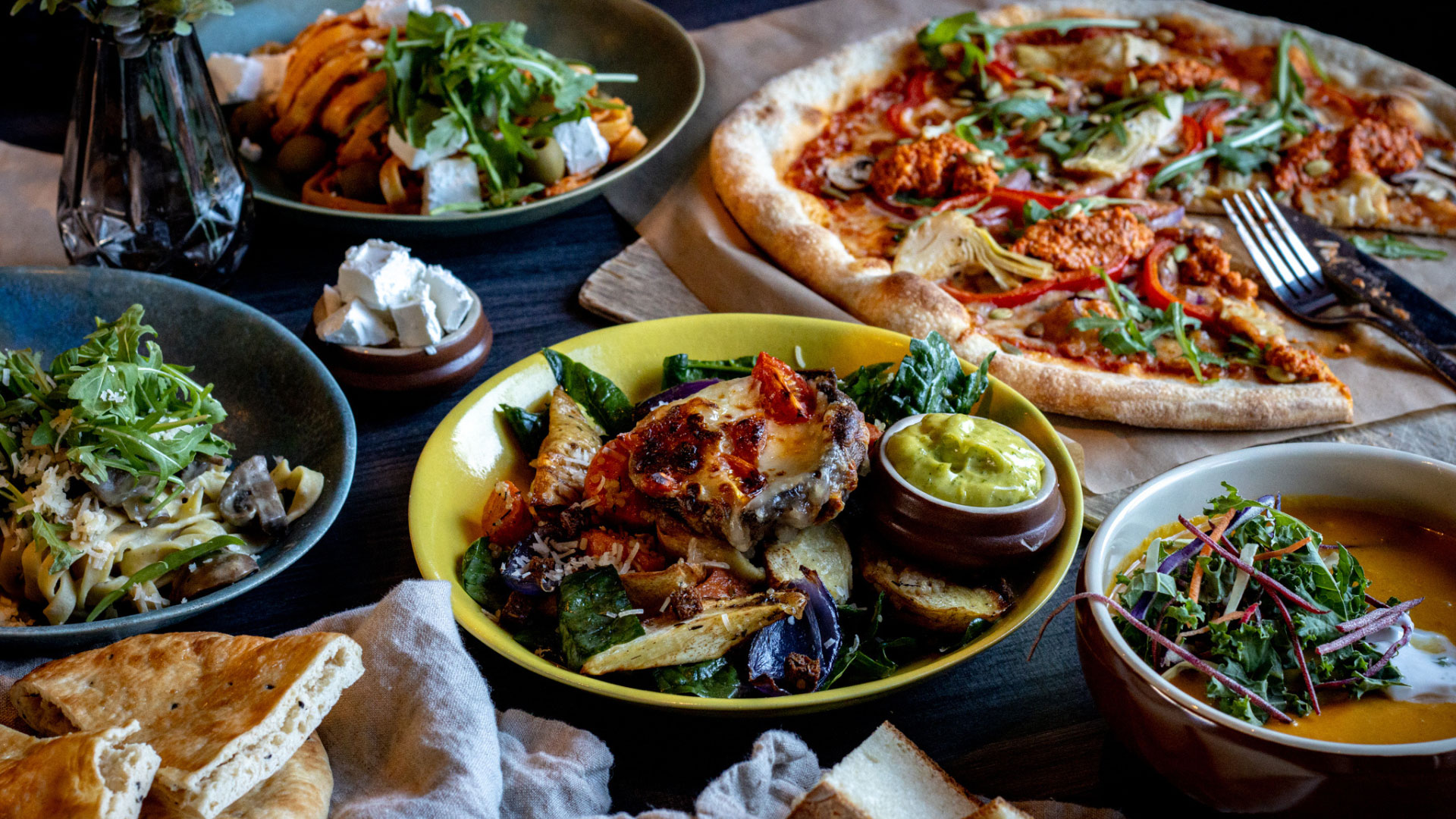 En mängd olika maträtter på ett bord, inklusive pizza, pastarätt med svamp, rostade grönsaker med sås och grönsallad