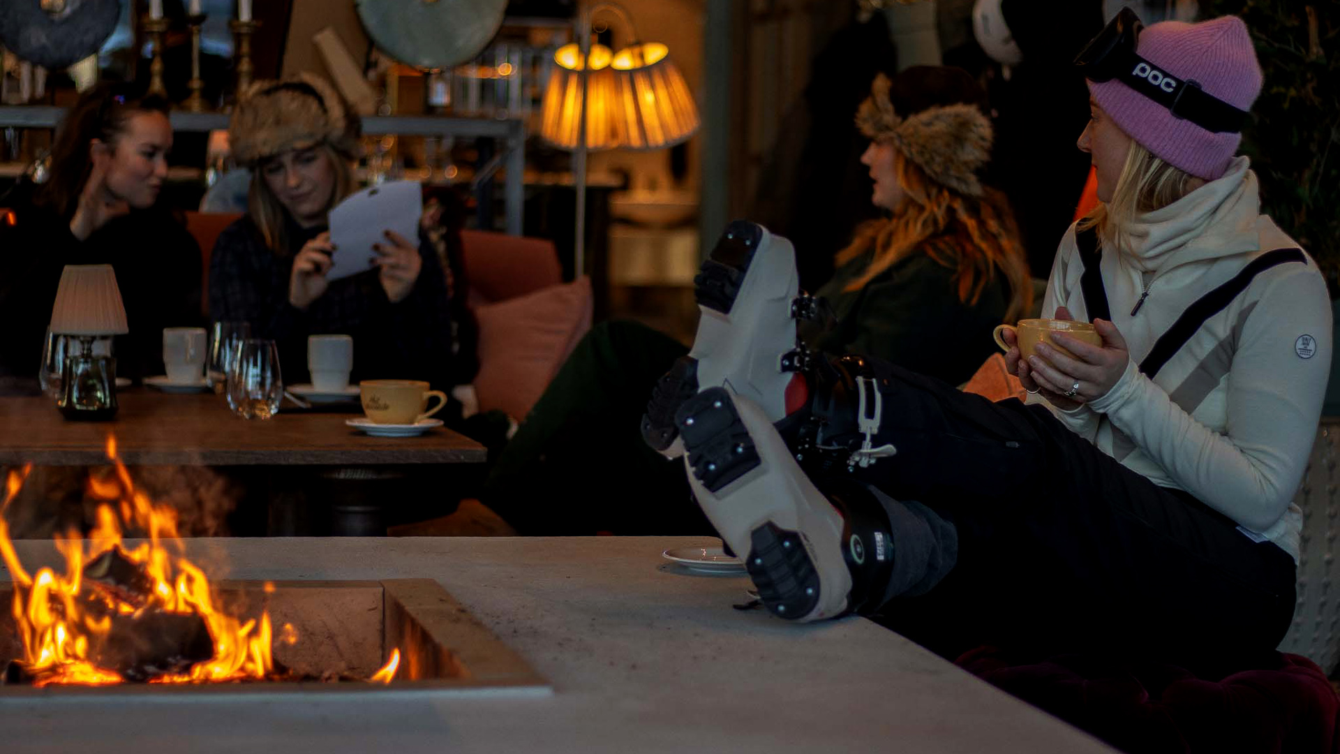 Personer kopplar av vid en brasa inomhus, iförda vinterkläder och skidutrustning, och dricker kaffe