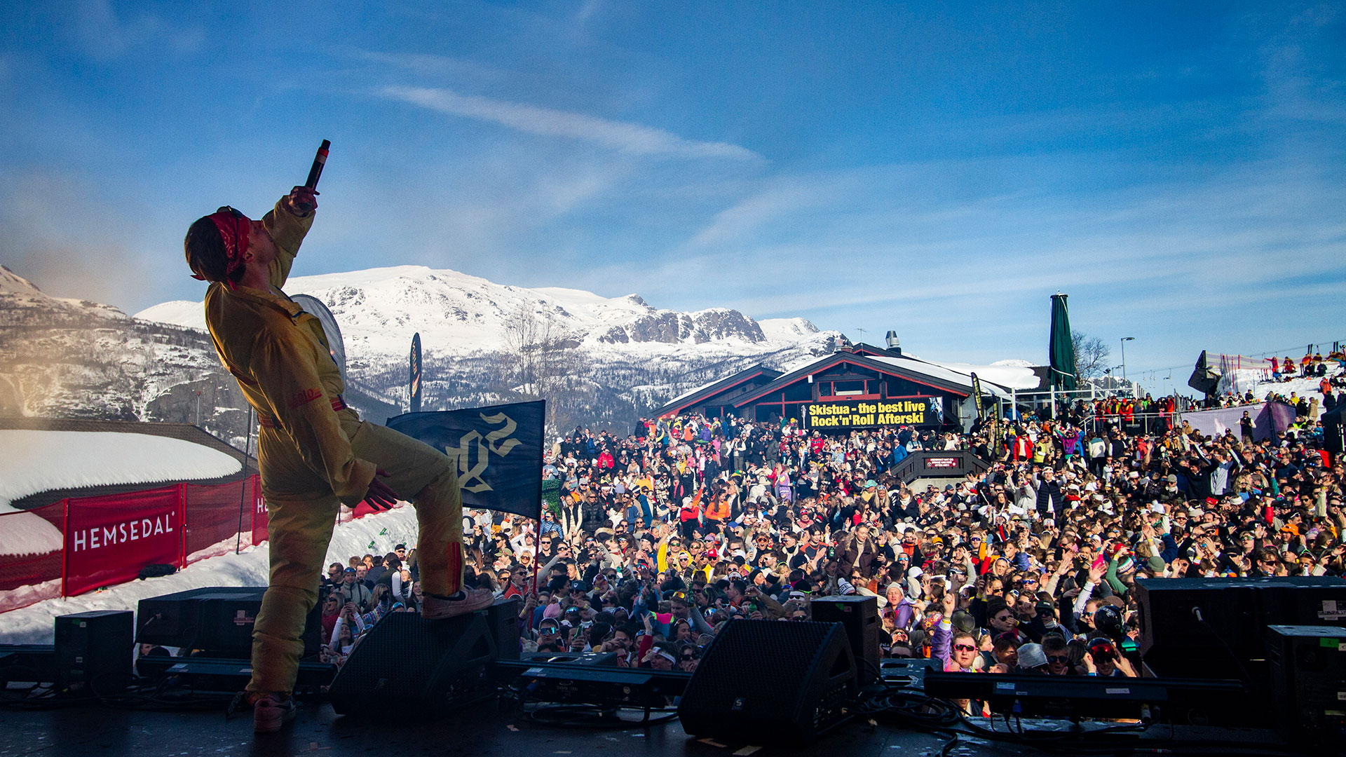 Artist uppträder på scen under livlig utomhuskonsert i Hemsedal med snötäckta fjäll i bakgrunden och stor publik i solskenet