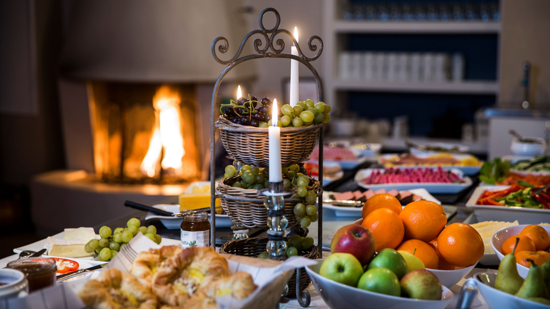 Lyxig frukostbuffé med färska frukter, bröd och pålägg i mysig miljö med brinnande eldstad i bakgrunden