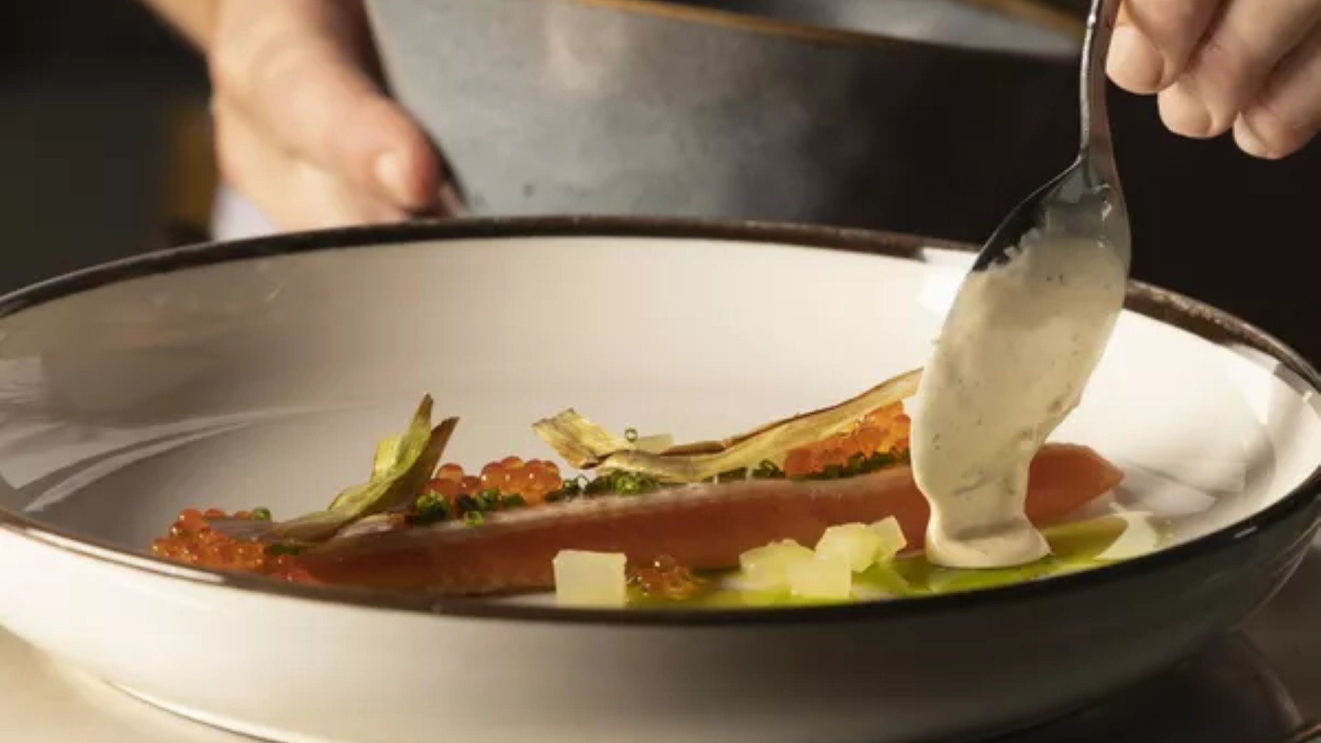 Kock garnerar en elegant tallrik med en skiva lax, dekorerad med röd kaviar och fänkål, genom att skeda en delikat sås över rätten