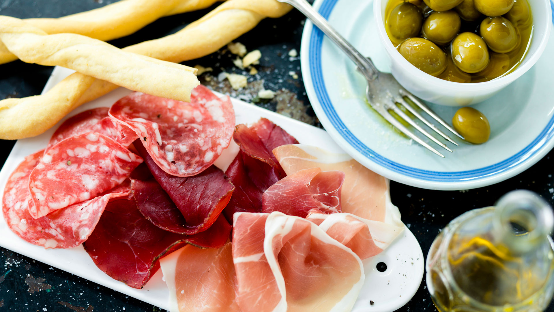 Charkbricka med olika sorter av salami, bresaola och parmaskinka, serverad med gröna oliver och krispiga brödpinnar