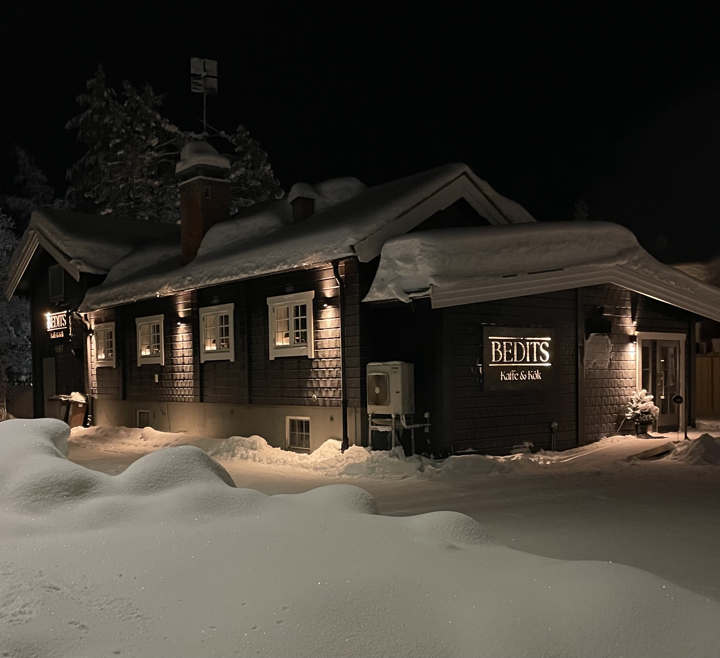 Snötäckt traditionell byggnad med skylt "Bedits Kaffe & Kök" upplyst i natten, omgiven av snödrivor
