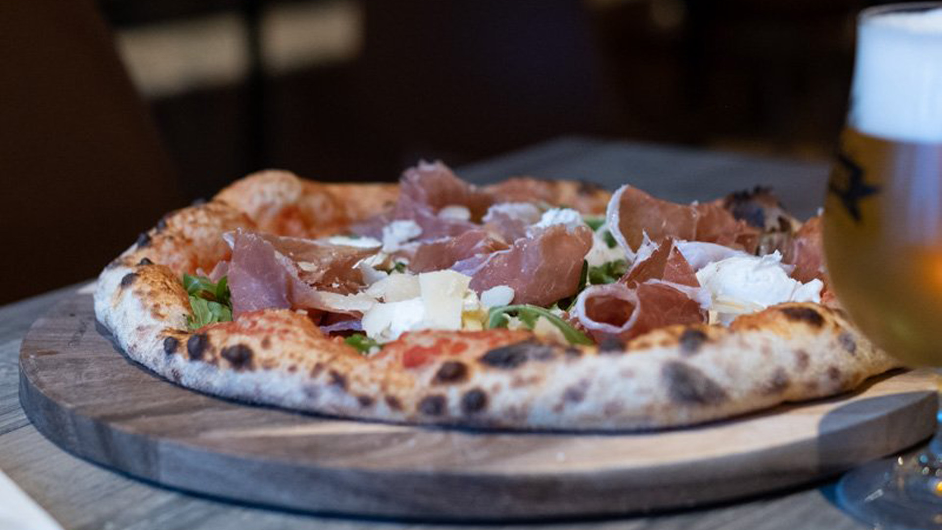 Pizza på träbricka toppad med prosciutto och parmesan, med ett glas öl i bakgrunden