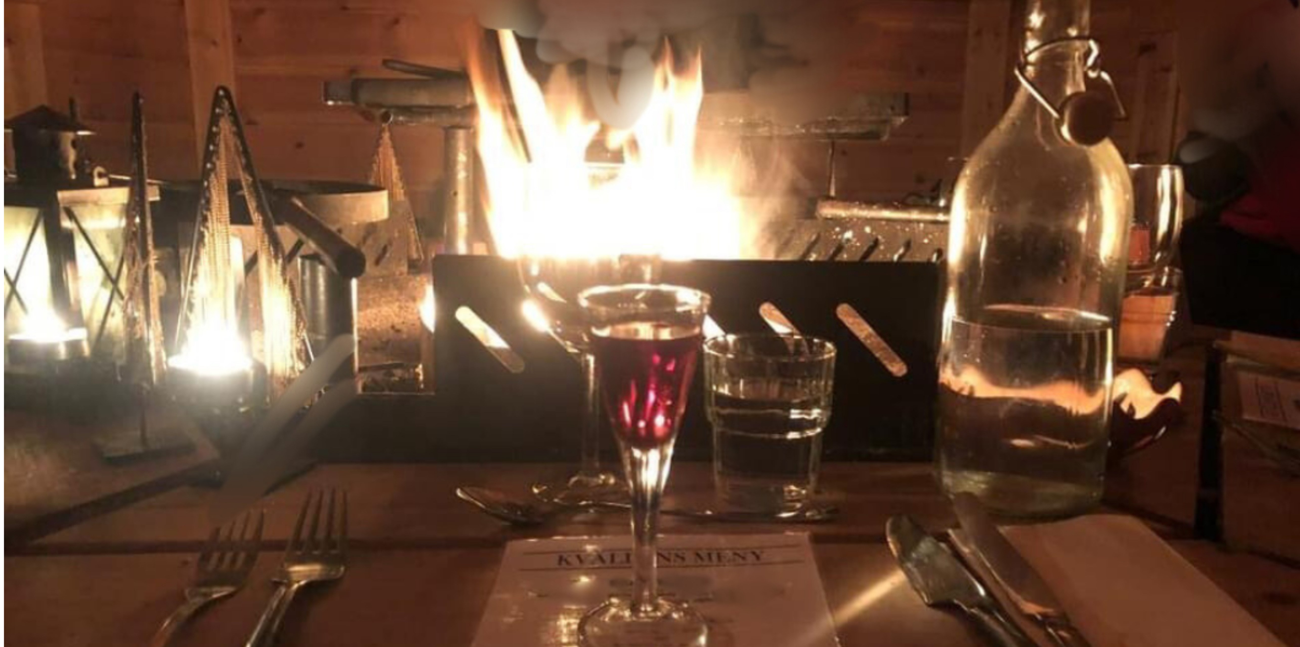 Rustik restaurangbordsinställning med en brinnande eld i bakgrunden, ett glas rött vin, vattenkaraff och menyer på träbord