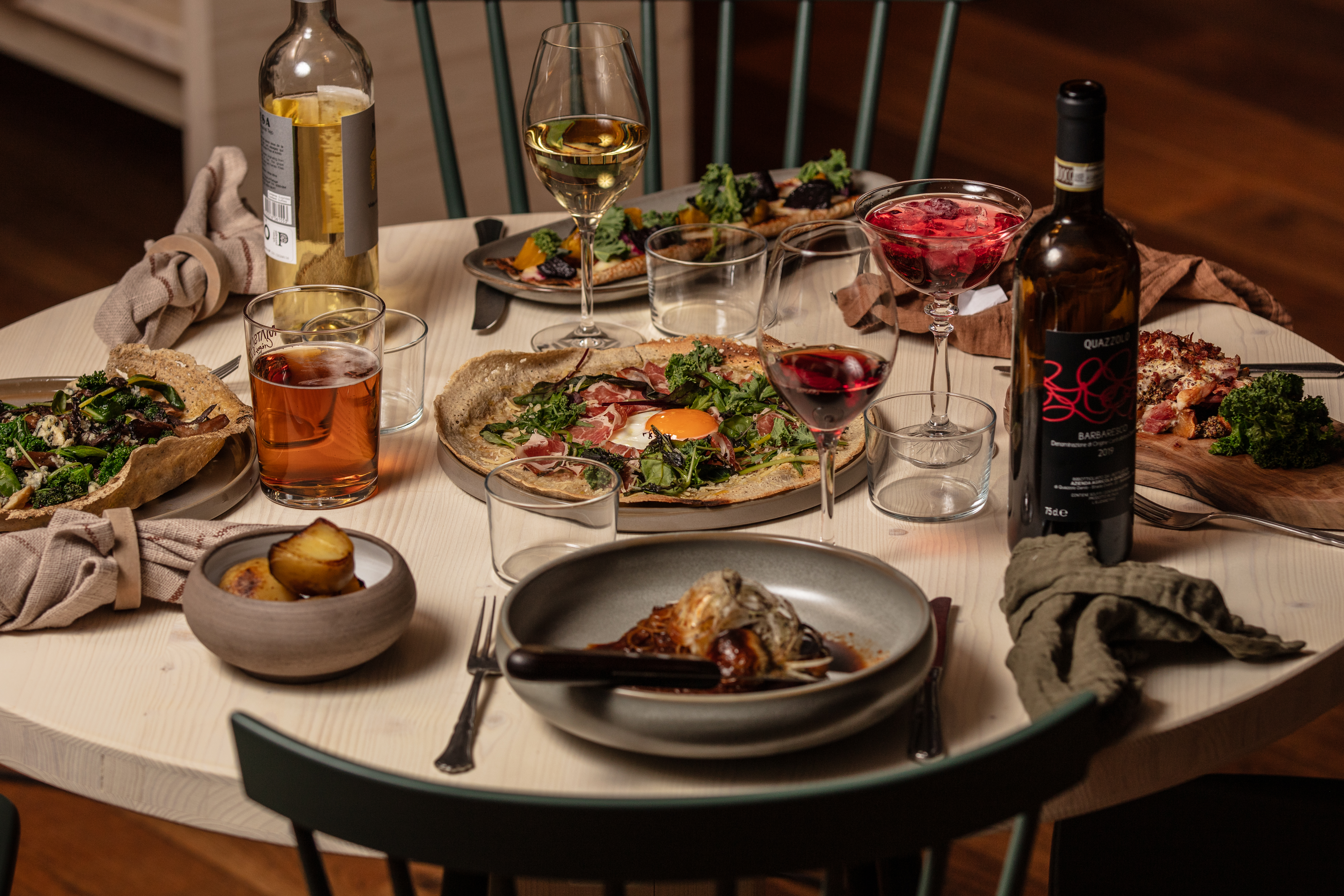 Middagsscen med olika rätter och drycker på ett runt bord, inklusive vin, öl och cocktails, samt tallrikar med mat som pizza och sallad