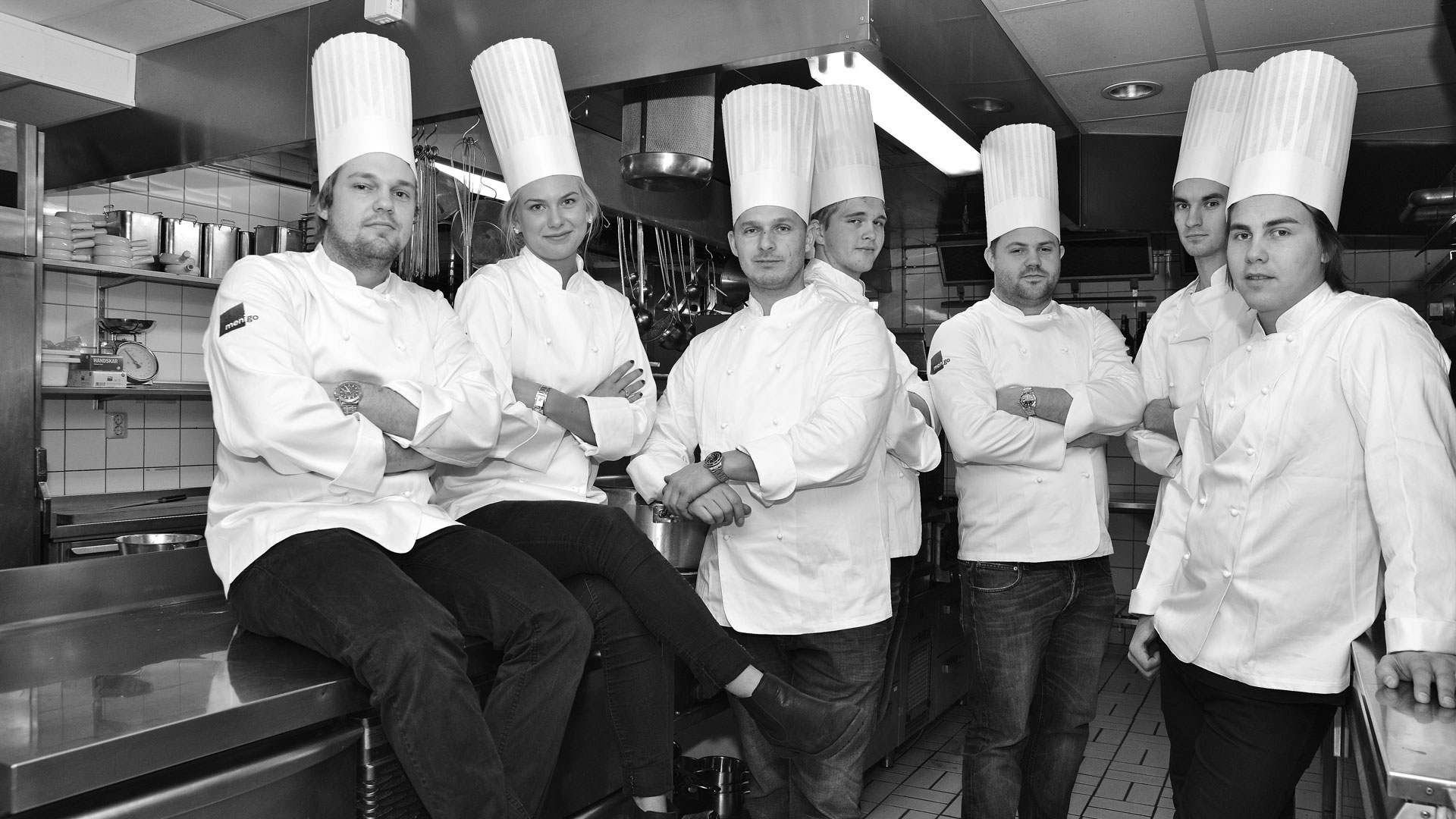Sju kockar i vita uniformer och höga kockmössor står i ett professionellt restaurangkök