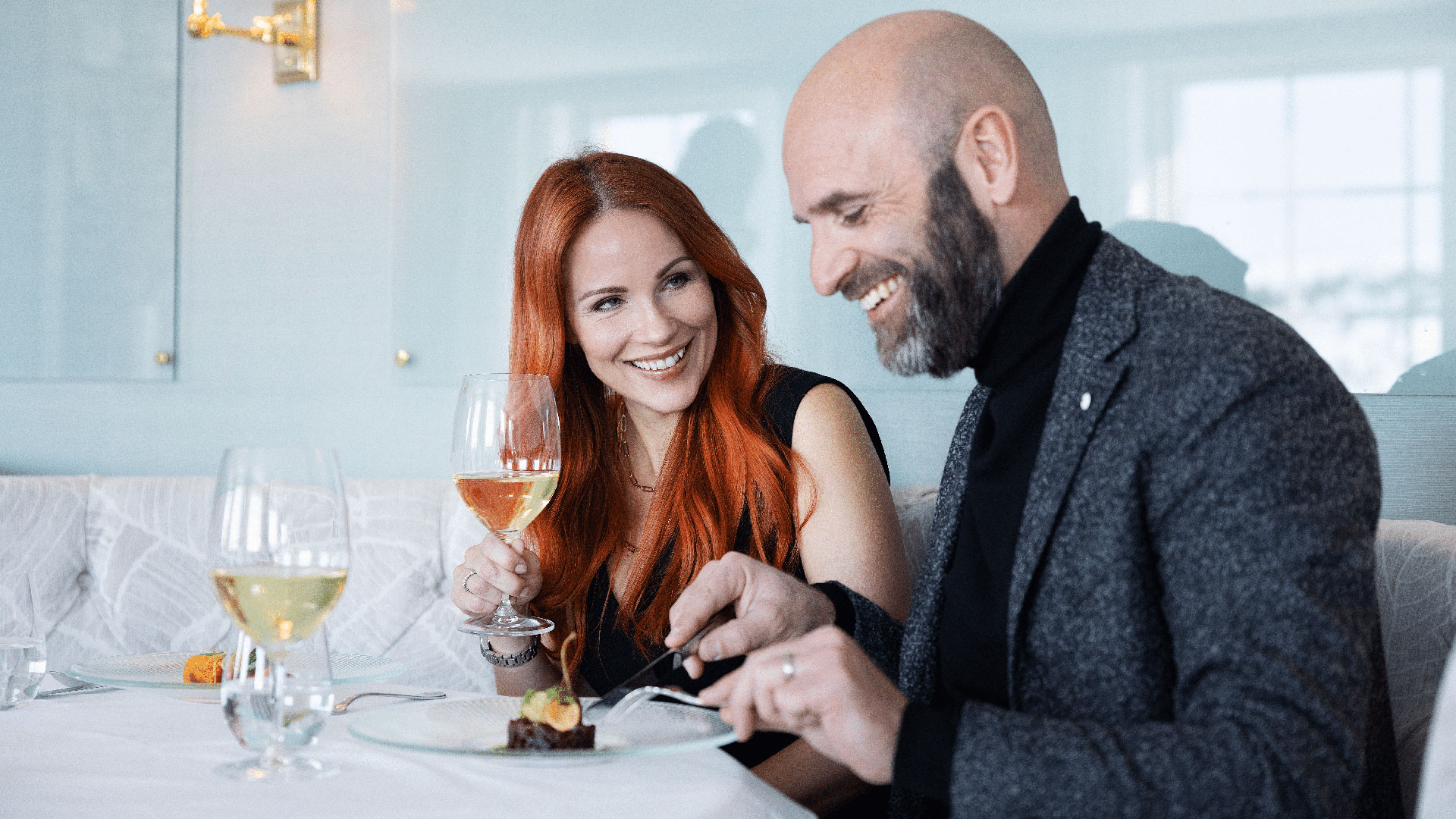 Par som njuter av en måltid på en restaurang, med varsitt glas vin och ler mot varandra i en ljus och elegant miljö