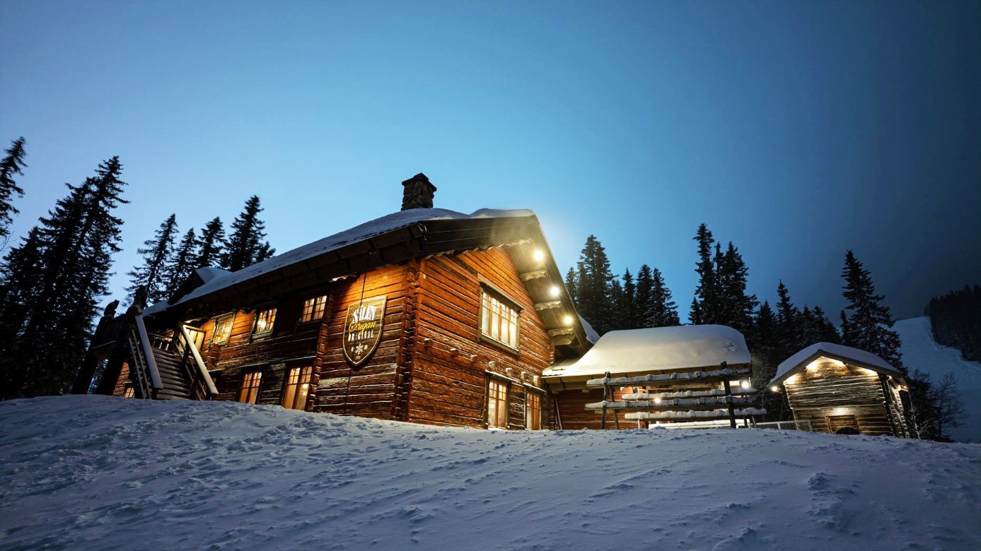Snötäckt timmerstuga i skogen vid skymning, upplyst med varma lampor, omgiven av granträd