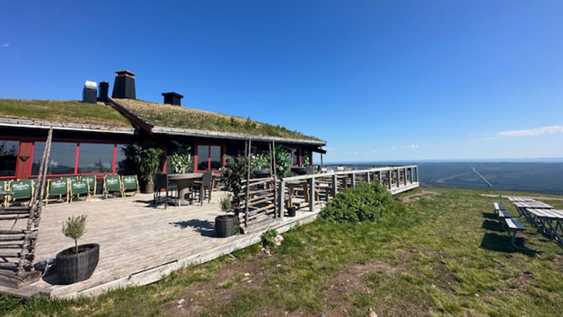 Restaurang med grästak på fjälltoppen, stor altan med utsikt över de vidsträckta skogarna under en klarblå himmel