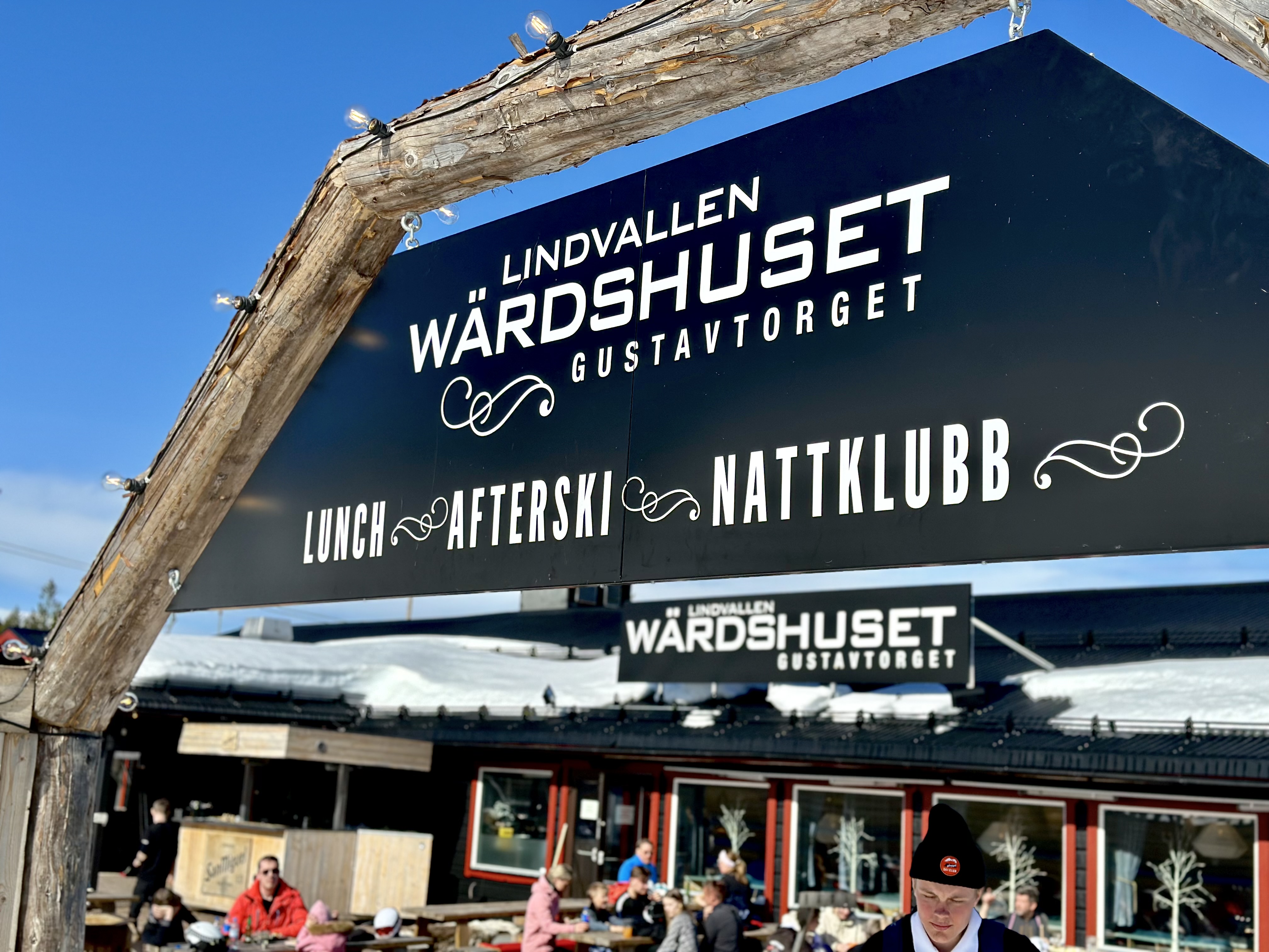 Skylt vid Lindvallen Wärdshuset Gustavtorget som annonserar lunch, afterski och nattklubb, med en rustik träram och vintermiljö i bakgrunden