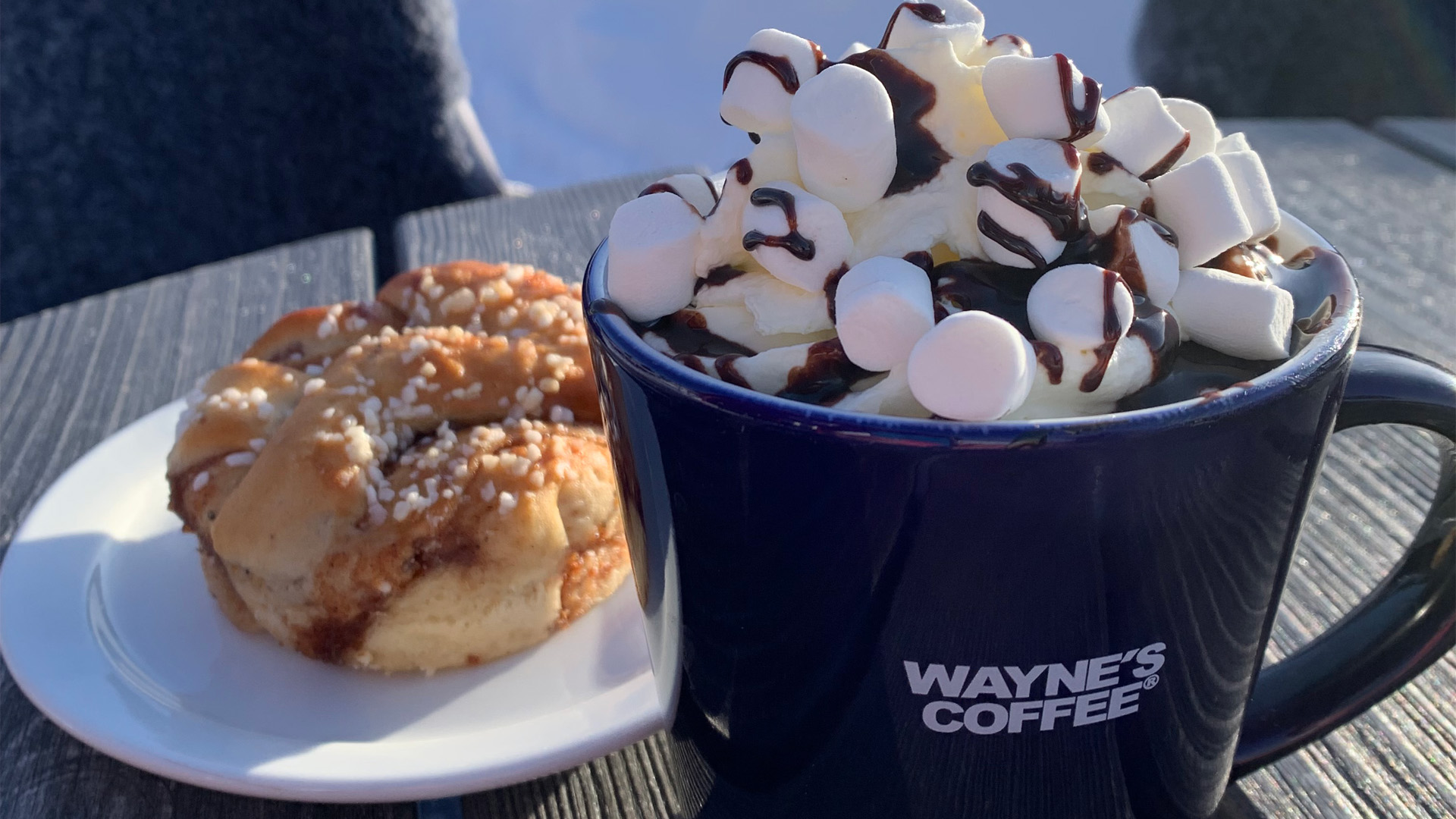 Chokladdryck med marshmallows i en mugg från Wayne's Coffee bredvid en kanelbulle på ett träbord
