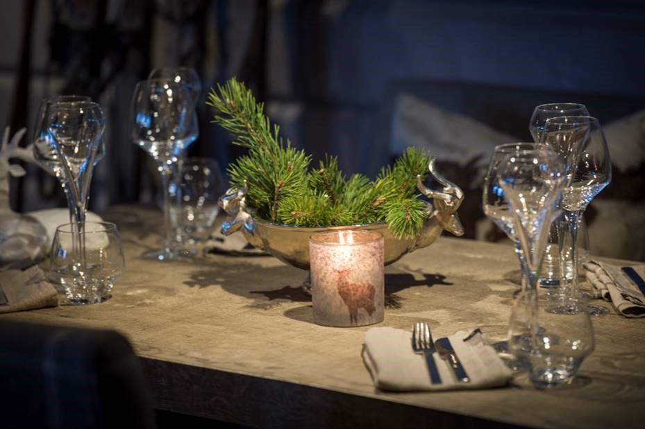 Festligt dukat bord med glas, servetter och grönt arrangemang i en dekorativ skål, belyst av ett stämningsfullt ljus