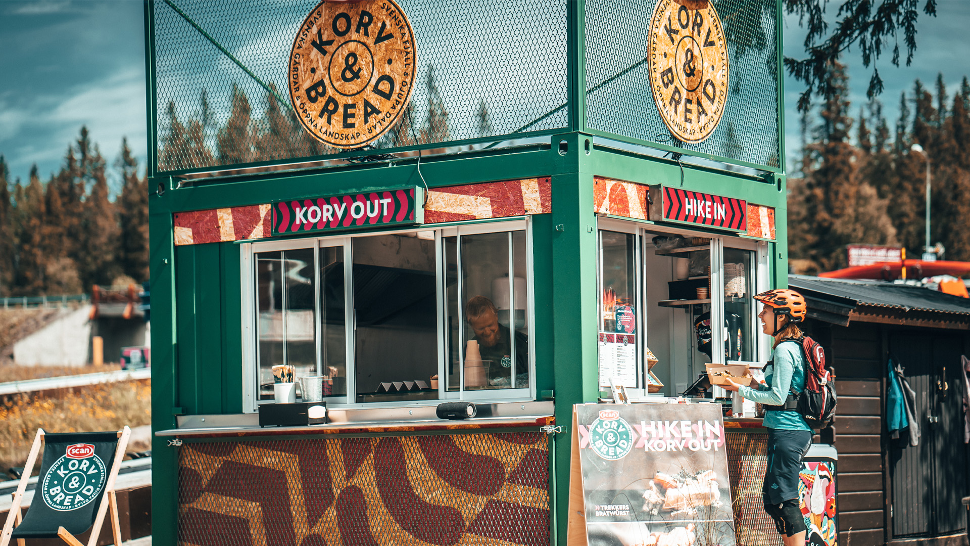Grön food truck med skylten "Korv & Bread" vid skogsområde, kund i cykelhjälm beställer mat