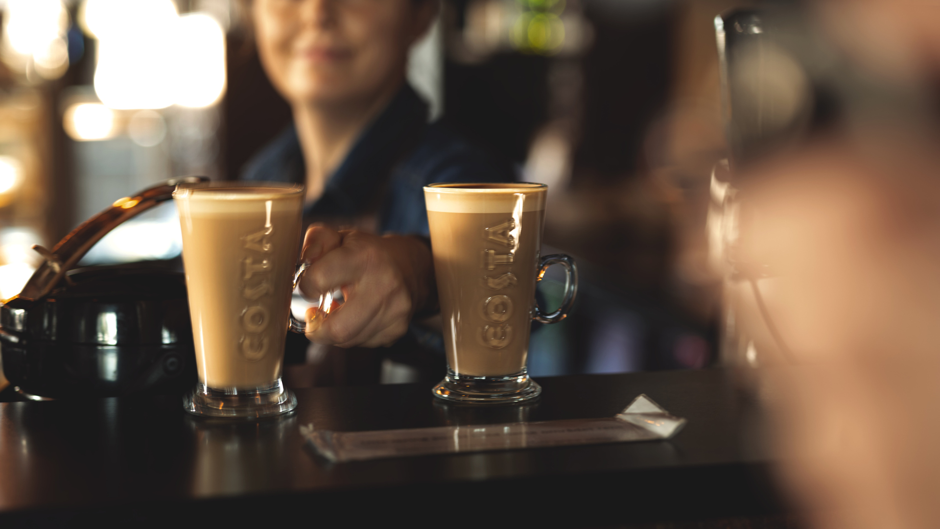 Två glas kaffe lattes på en kafédisk med suddig bakgrund, en person räcker fram ett glas