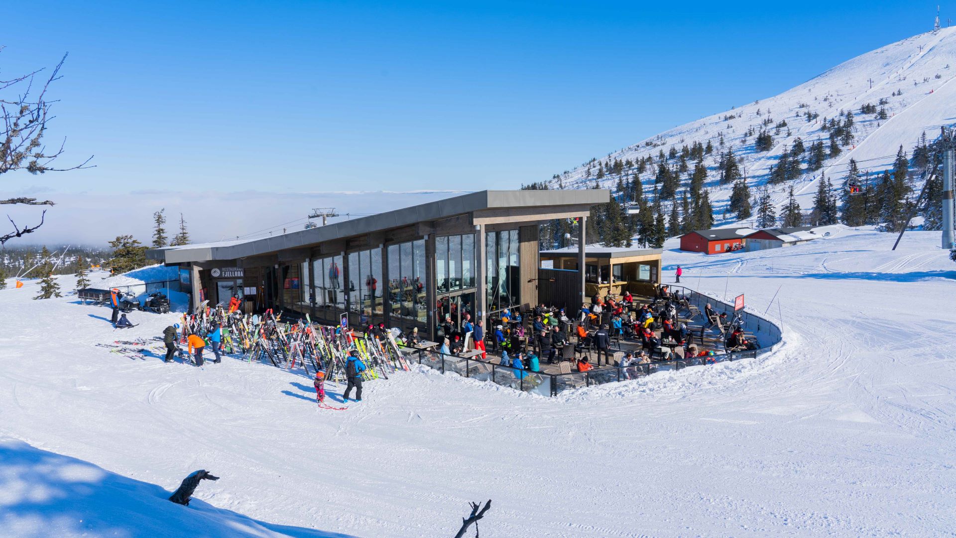 Skidåkare samlade vid fjällrestaurang med snötäckta berg och klarblå himmel i bakgrunden