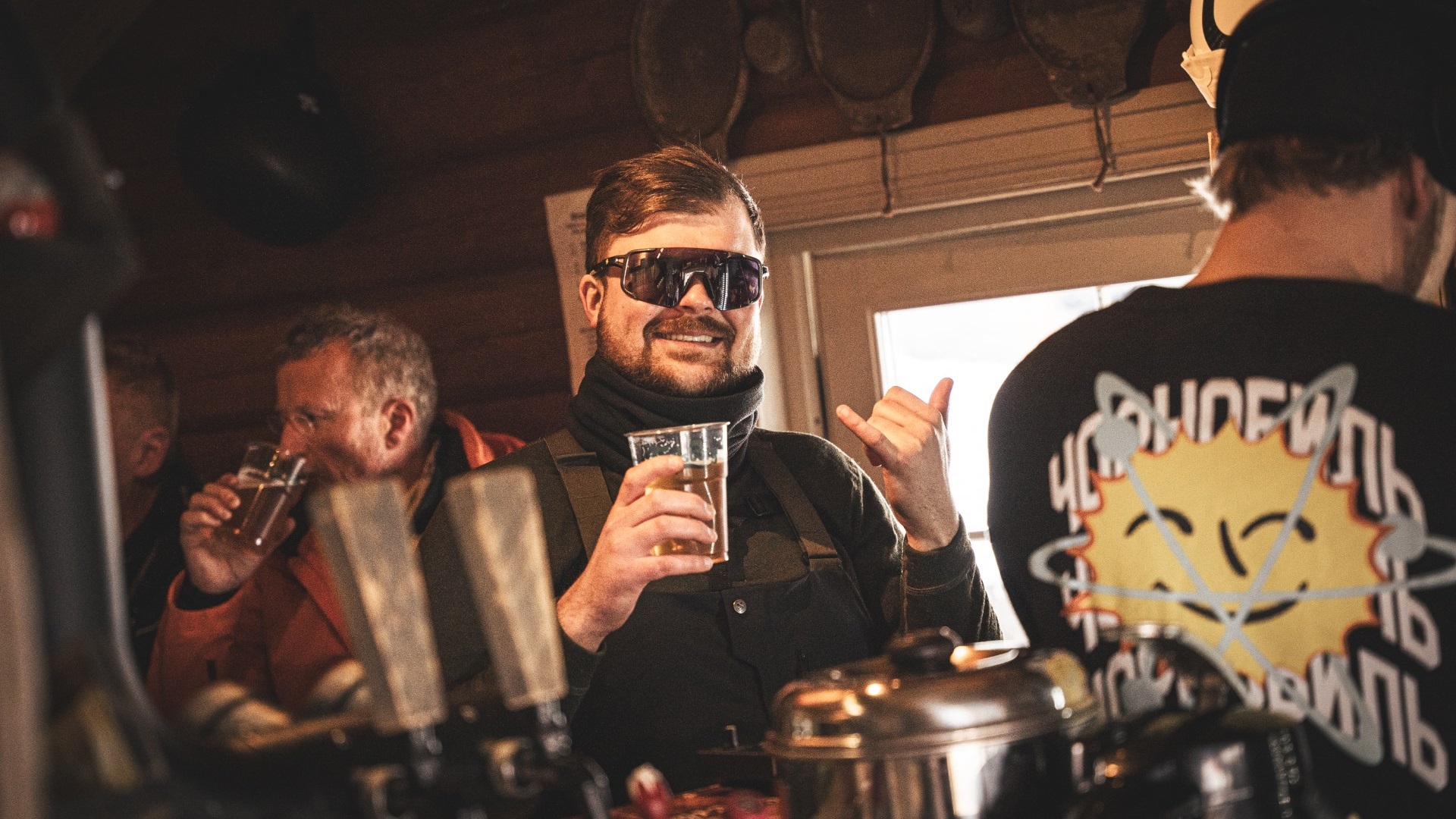 En man med solglasögon och skägg håller en öl på en livlig bar. Andra personer i bakgrunden pratar och dricker, skapar en avslappnad atmosfär