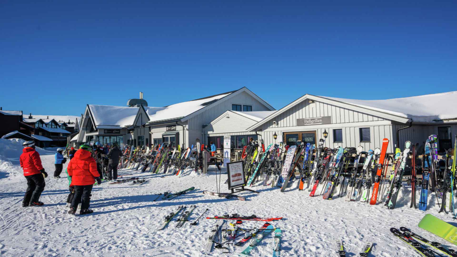 Skiduthyrning och aktiviteter vid ett vintersportcenter med snötäckta byggnader och uppställda skidor, personer i färgglada kläder förbereder sig för skidåkning under klarblå himmel
