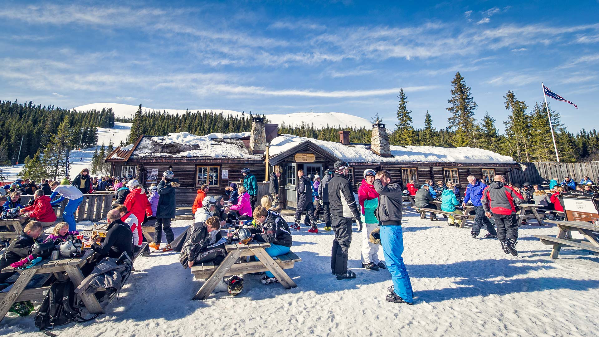 Knettsetra i Trysil och besökare i färgglada kläder, sitter vid träbord och njuter av solen. Vinterlandskap med snötäckta fjäll och skog i bakgrunden