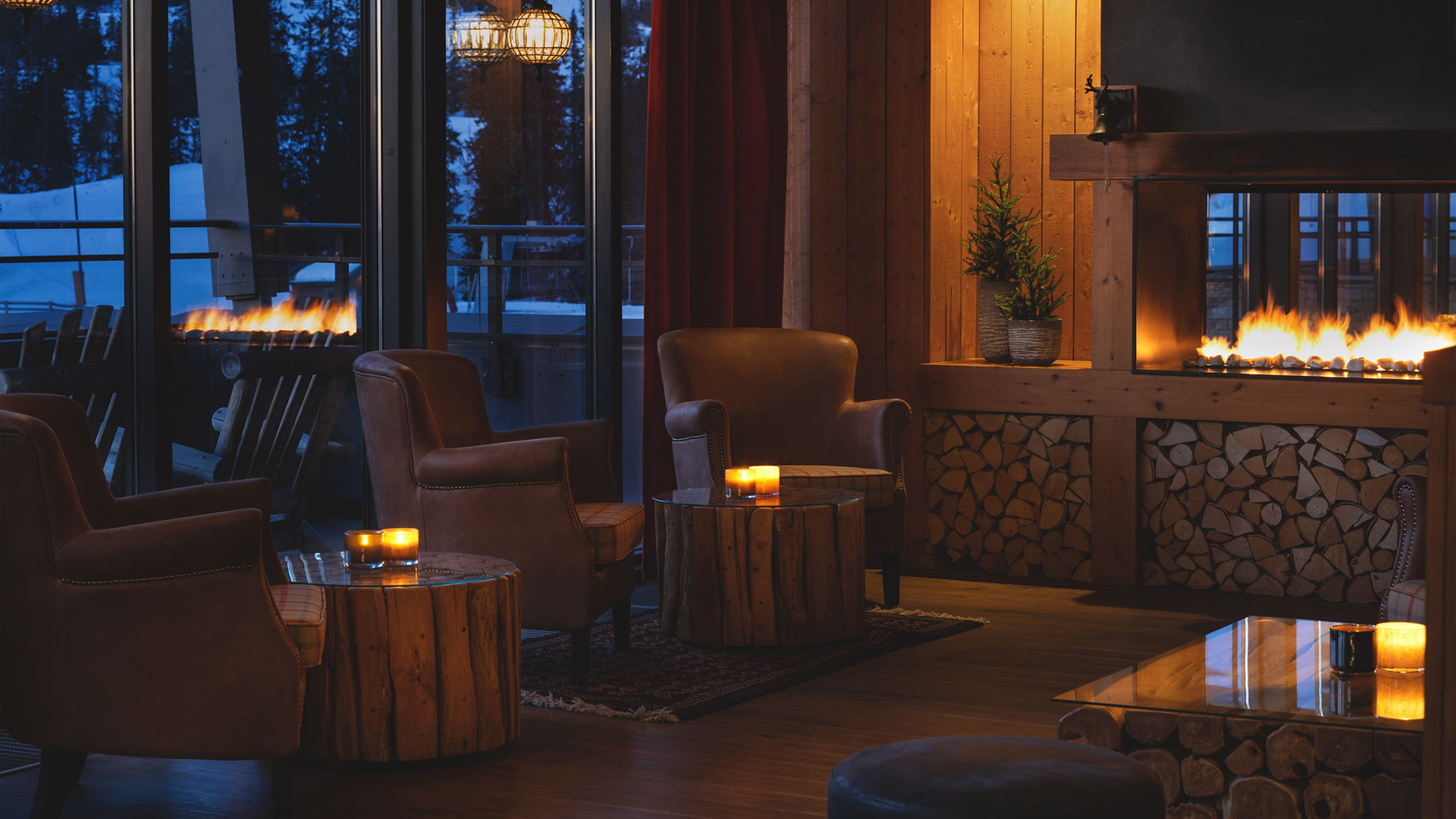 Mysig hotellobby med öppen spis, bekväma fåtöljer och ljus på runda träbord. Utsikt mot mörk skog och snötäckt terrass