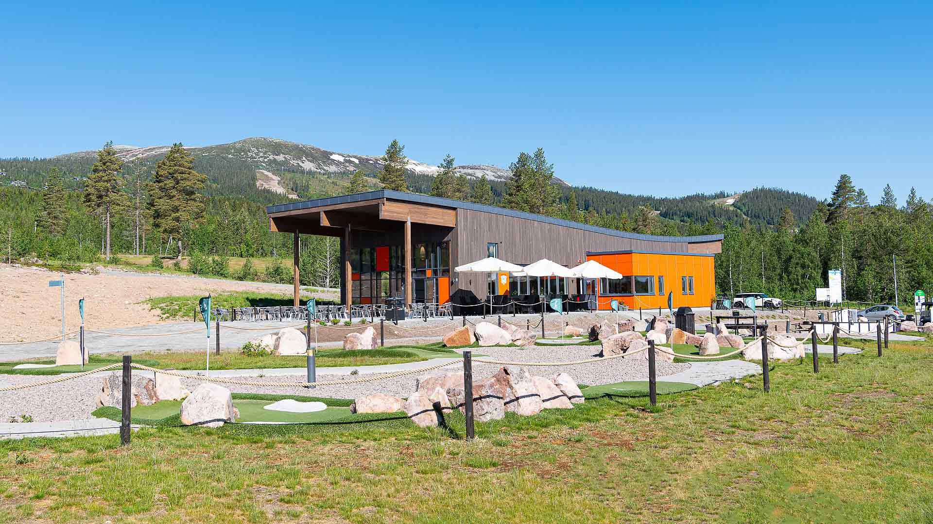 Modern byggnad i fjällmiljö med stor terrass och utsikt över grönskande landskap och berg i bakgrunden