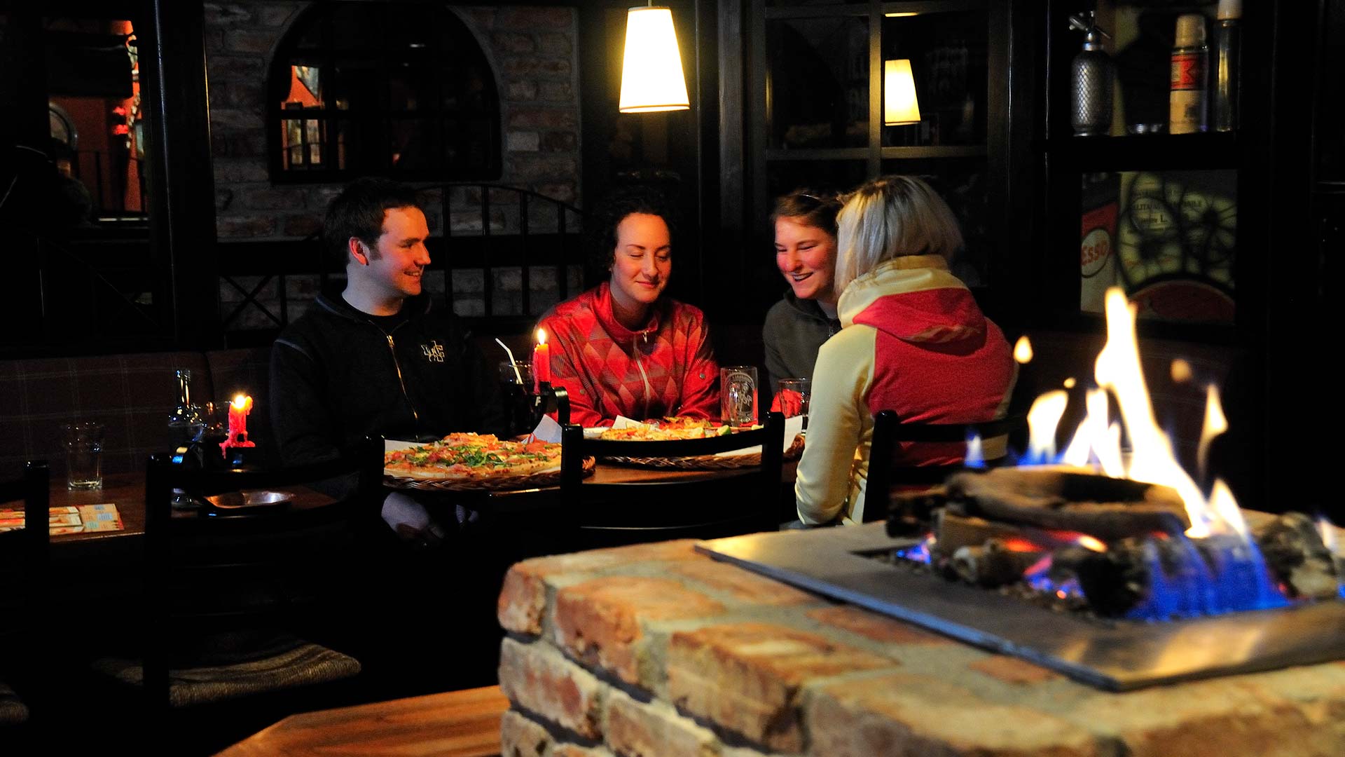Personer som njuter av pizza runt ett bord i en mysig restaurang med öppen spis i förgrunden