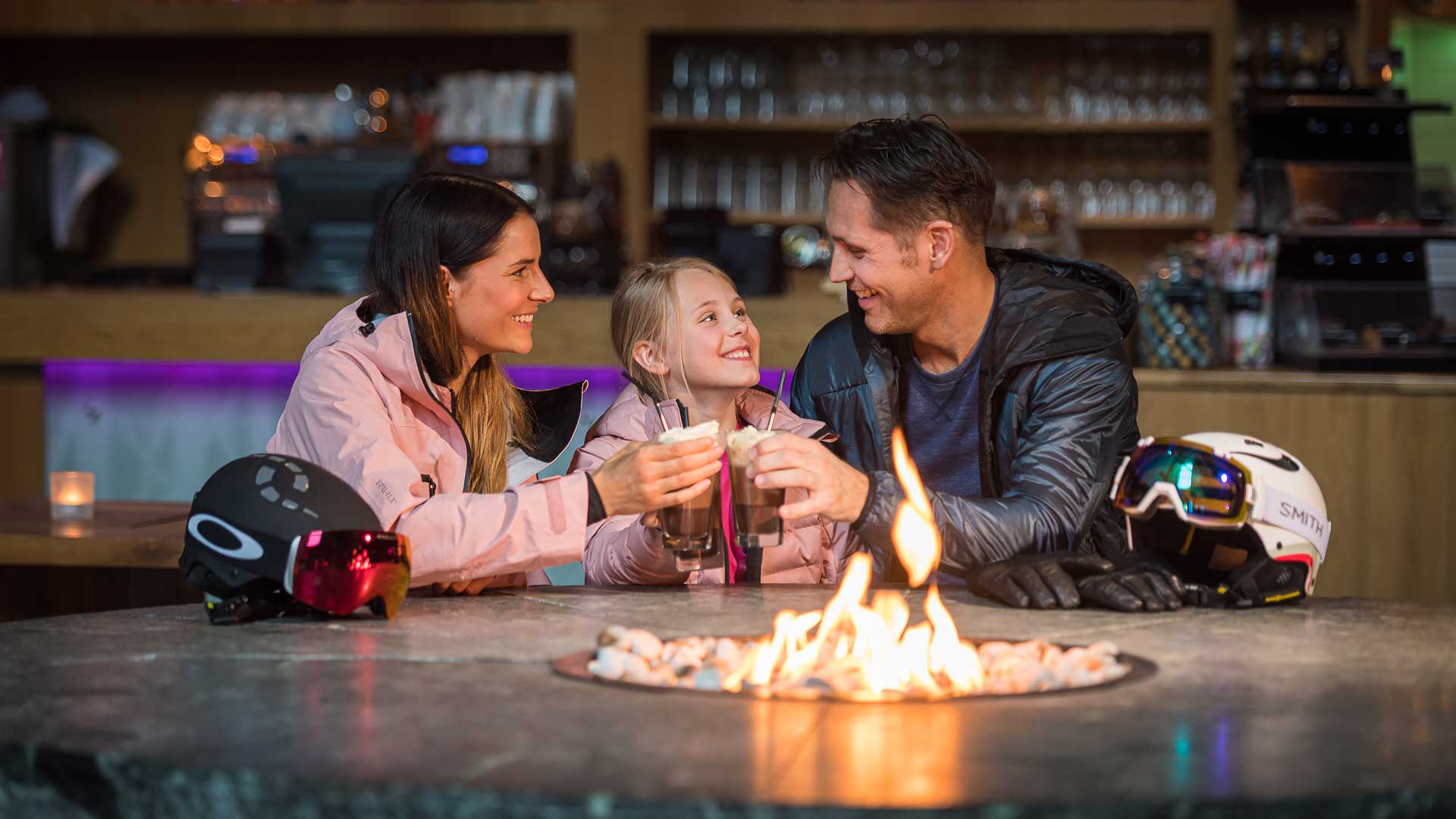 Familj umgås framför en öppen eld efter skidåkning, med hjälmar och skidglasögon på bordet