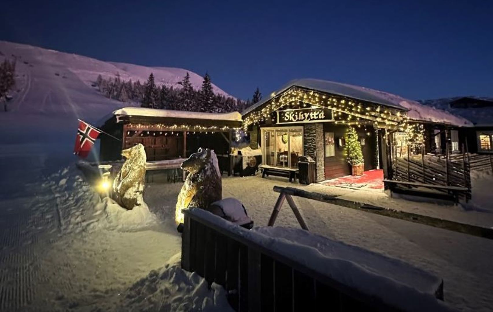 Snötäckt skidstuga i Norge dekorerad med julbelysning, norsk flagga och björnstatyer utanför i skymningen