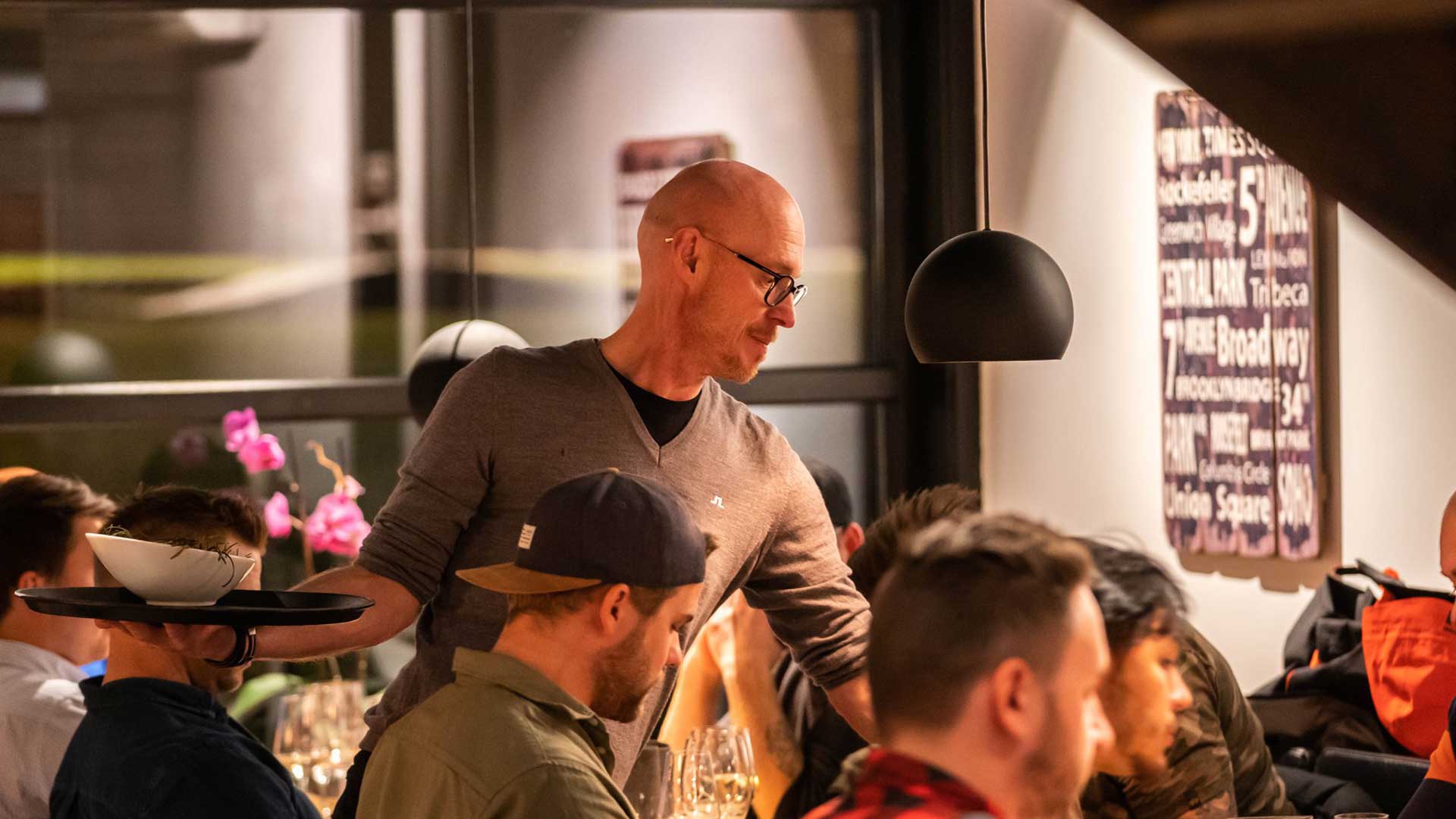 Person som serverar mat på en livlig restaurangmiljö, omgiven av gäster vid borden