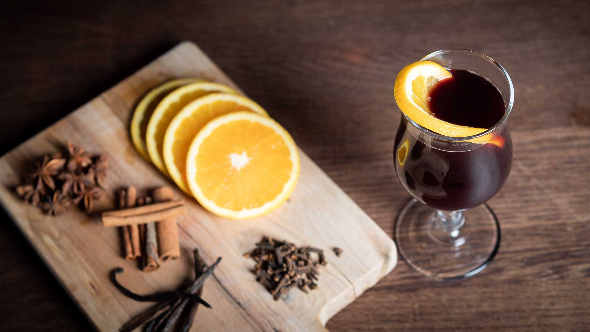 Glas med glögg dekorerat med apelsinskiva på ett träbord, omgiven av apelsinskivor, kanelstänger och stjärnanis