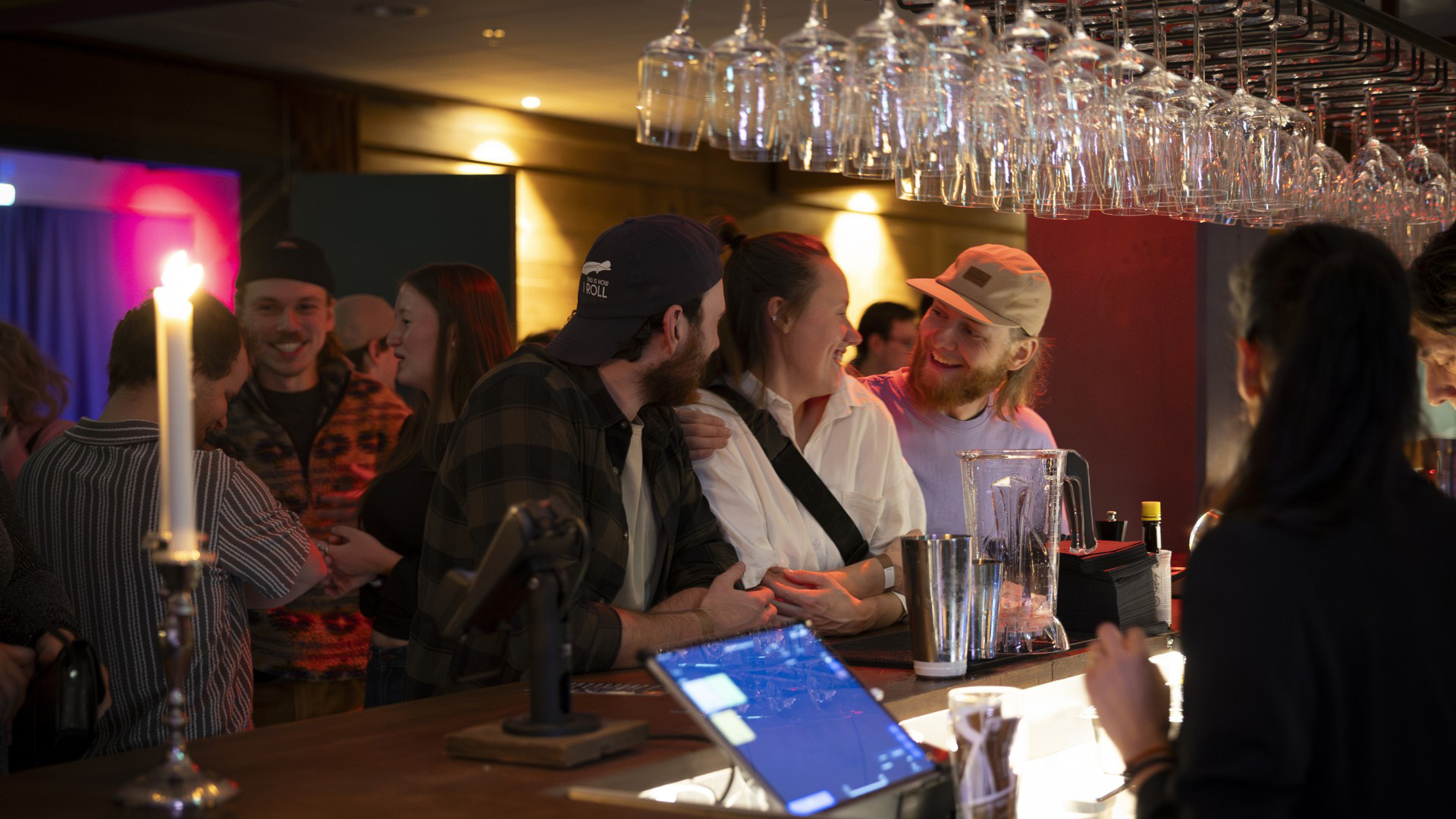 Personer socialiserar vid en bar med glas ovanför och levande ljus, i en varm och upplyst miljö