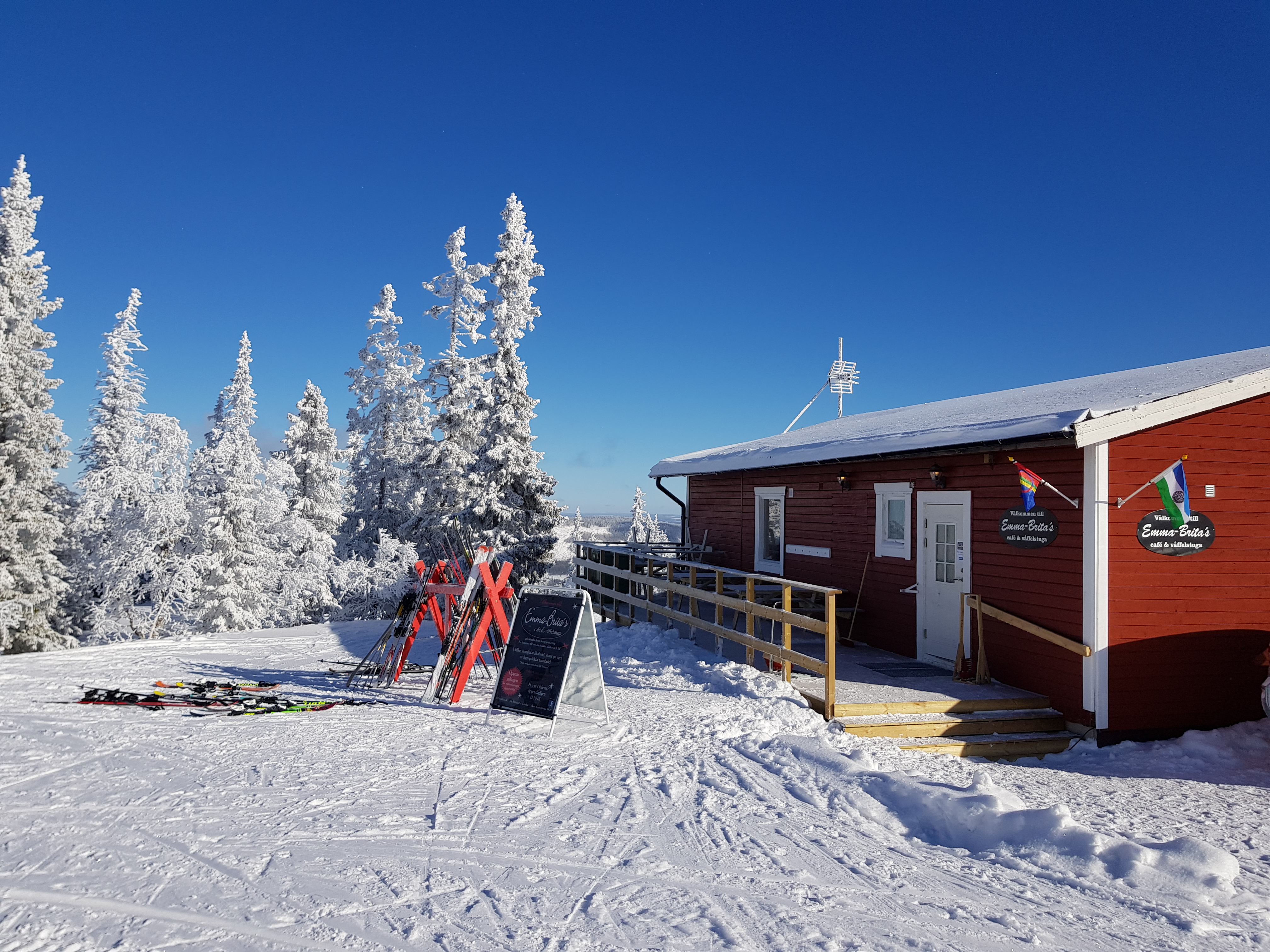 Röd stuga i vintrigt landskap med skidor uppställda utanför, omgiven av snötäckta träd och klarblå himmel. Perfekt för skidåkning i Sverige