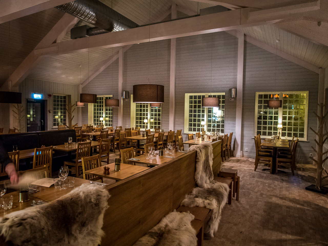 Restauranginteriör med träbord och stolar, mysbelysning och fårskinn på bänkarna, vilket skapar en varm och inbjudande atmosfär