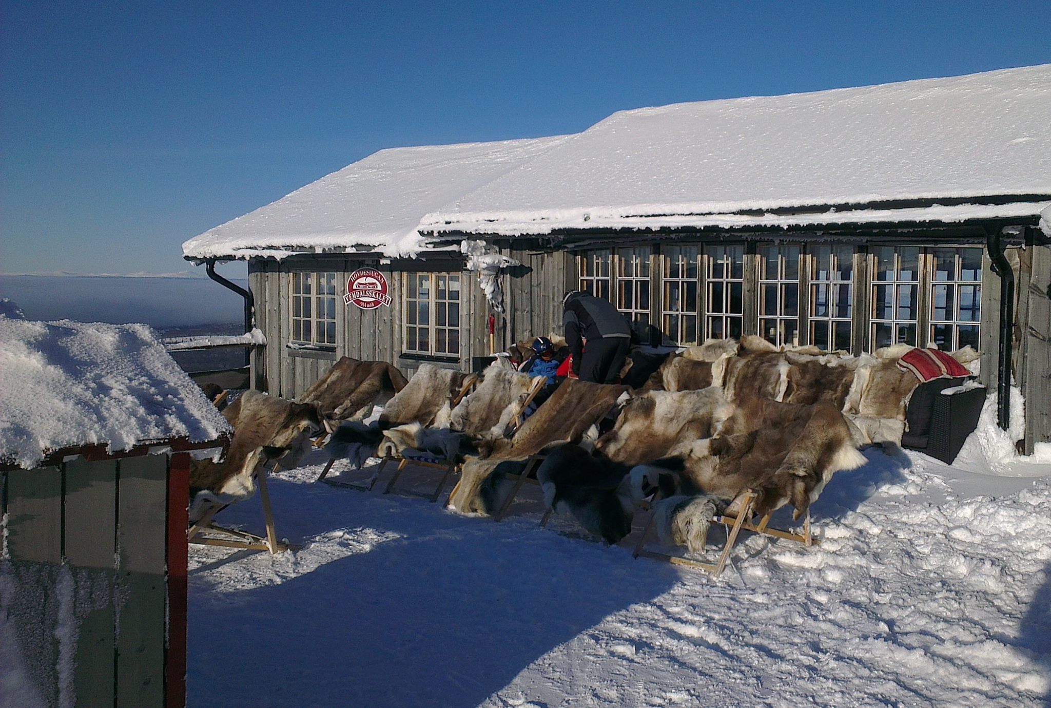 Snöig fjällstuga med renfällar på utestolar under solig vinterdag i Sverige