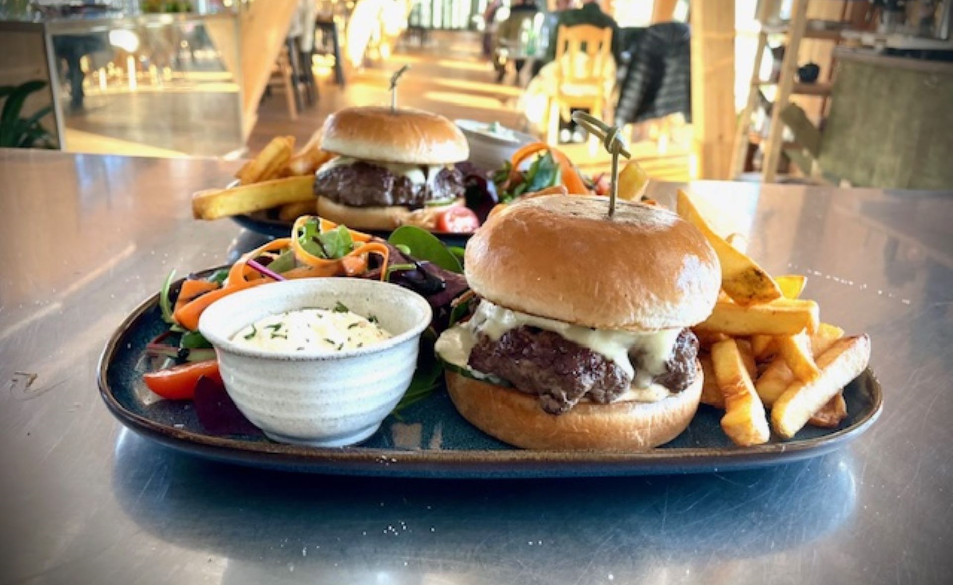 Två gourmetburgare med ost och pommes frites serveras på ett bord i en restaurangmiljö, åtföljda av färsk sallad och sås