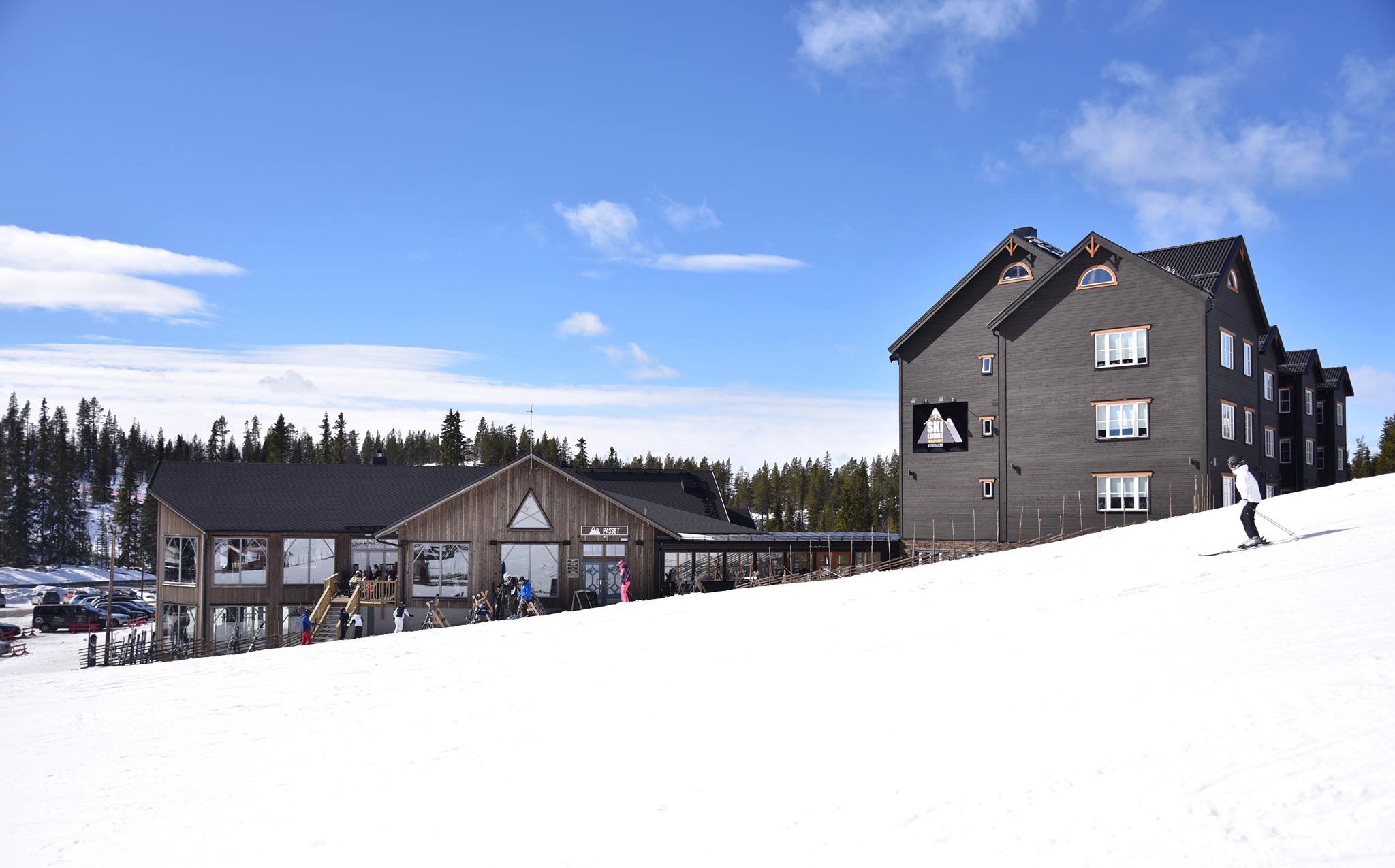 Skidåkare i en solig backe nära en fjällstuga i vinterlandskap, med snötäckta träd och klar blå himmel i bakgrunden