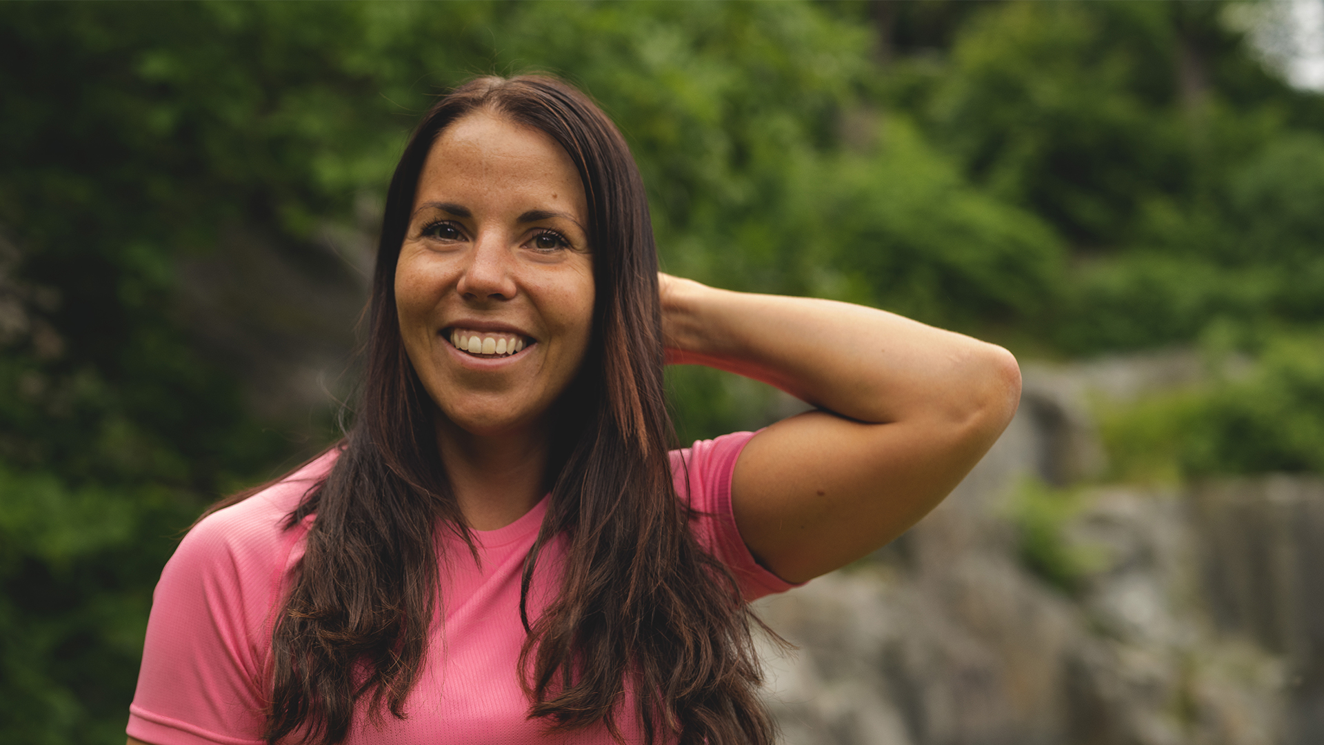 Kvinna i rosa t-shirt ler utomhus, omgiven av grönskande natur