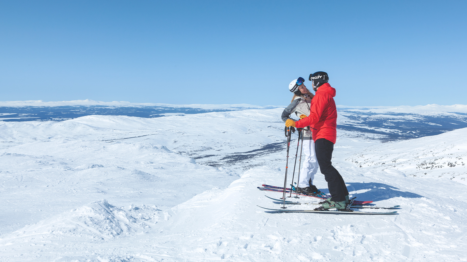Två skidåkare på snötäckt bergstopp i soligt väder med vidsträckt utsikt över fjällandskap