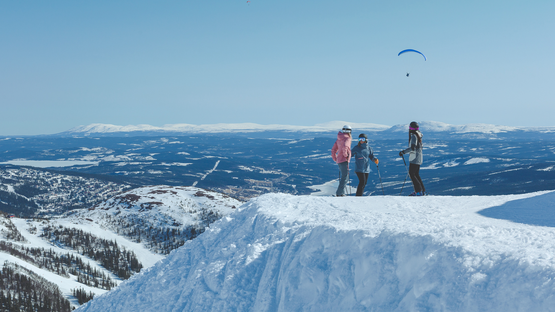 Tre personer på skidresa i fjällen med vidsträckt snölandskap och en paraglider i bakgrunden